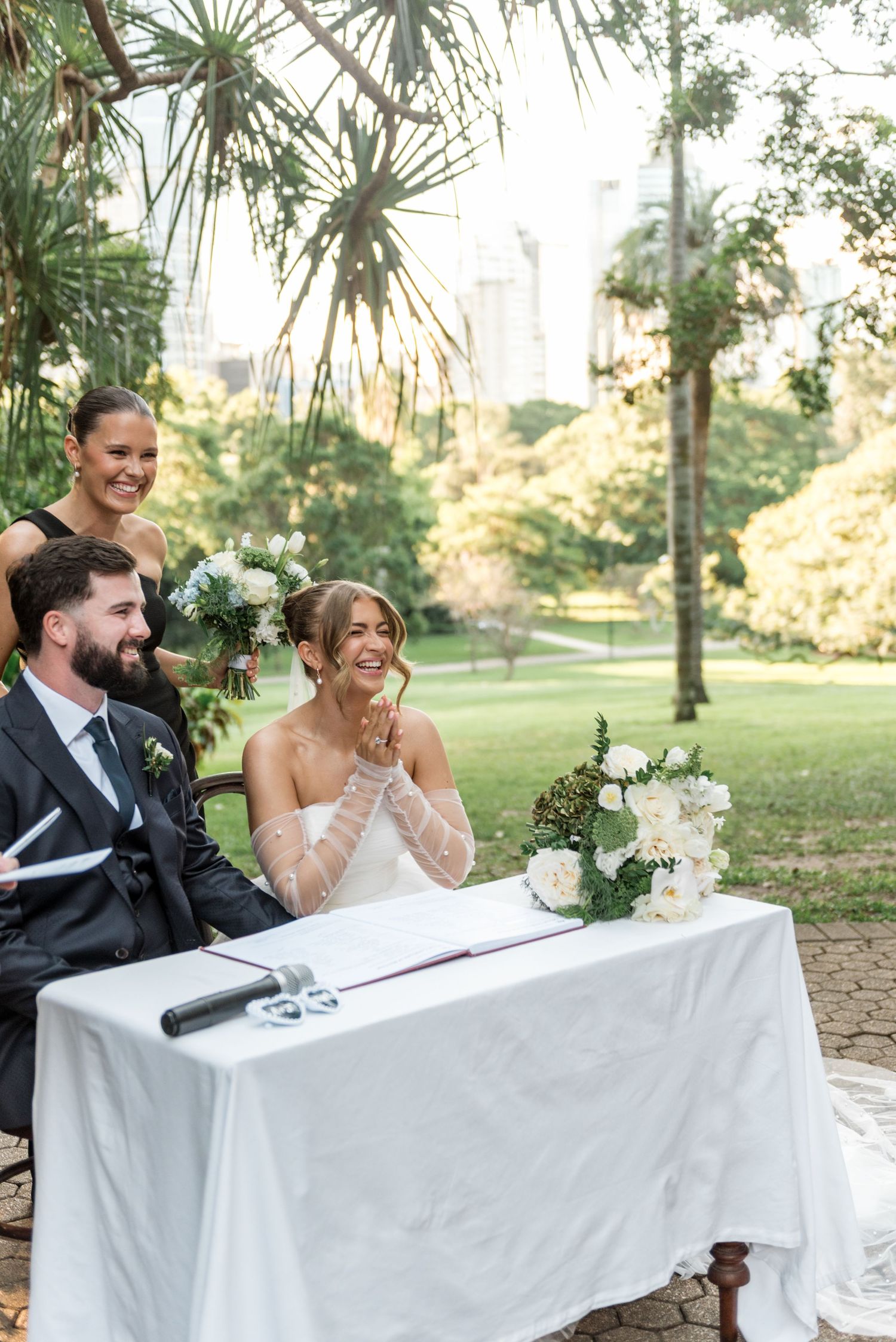 Bride and groom smile joyfully while signing wedding register at outdoor ceremony table with white flowers.