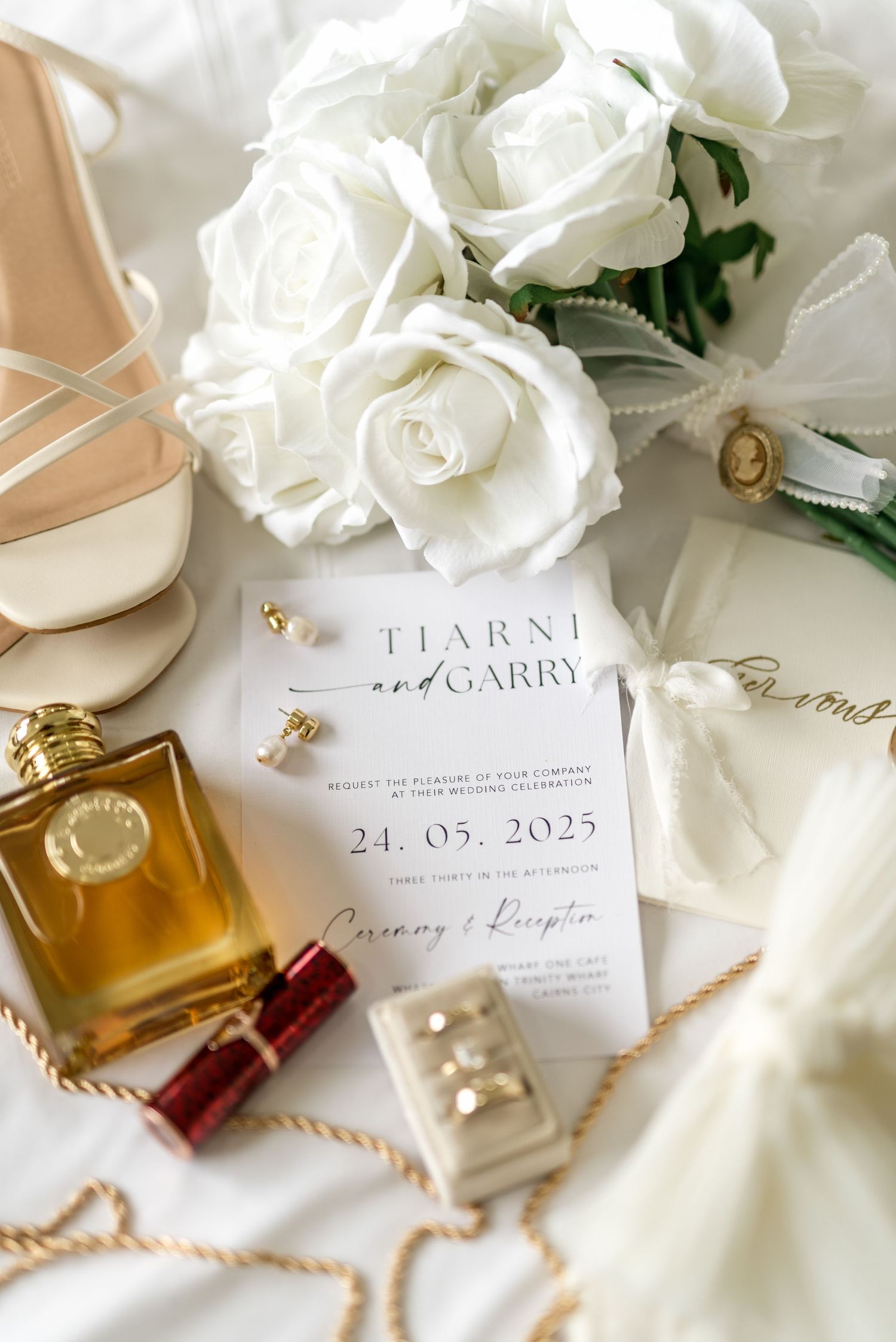 Wedding flat lay featuring Tiarni and Garry invitation dated 24.05.2025, white roses, nude heels, gold perfume, and jewelry.