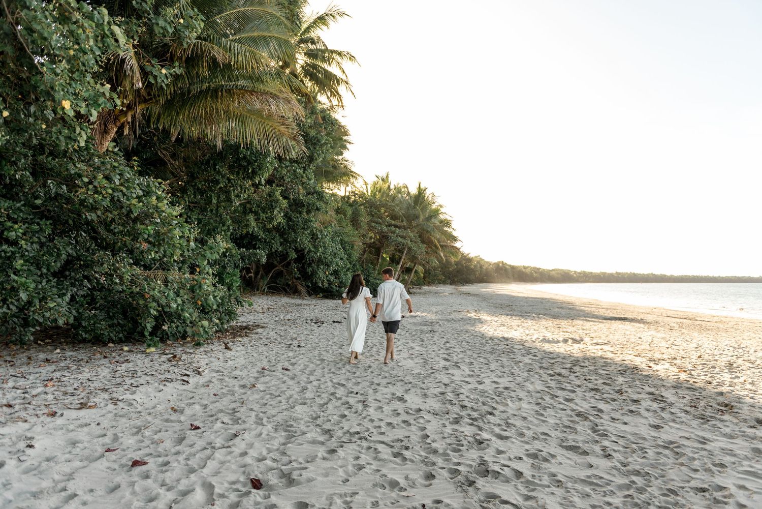 Couple walking hand in hand along a sandy tropical beach at golden sunset near palm trees.