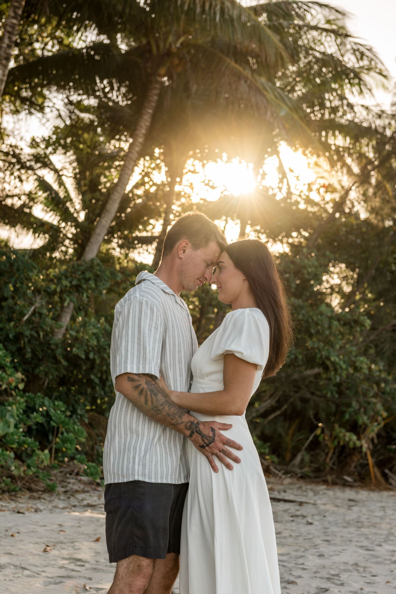 Romantic couple embracing forehead-to-forehead on a beach at sunset with tropical palm trees glowing behind them.