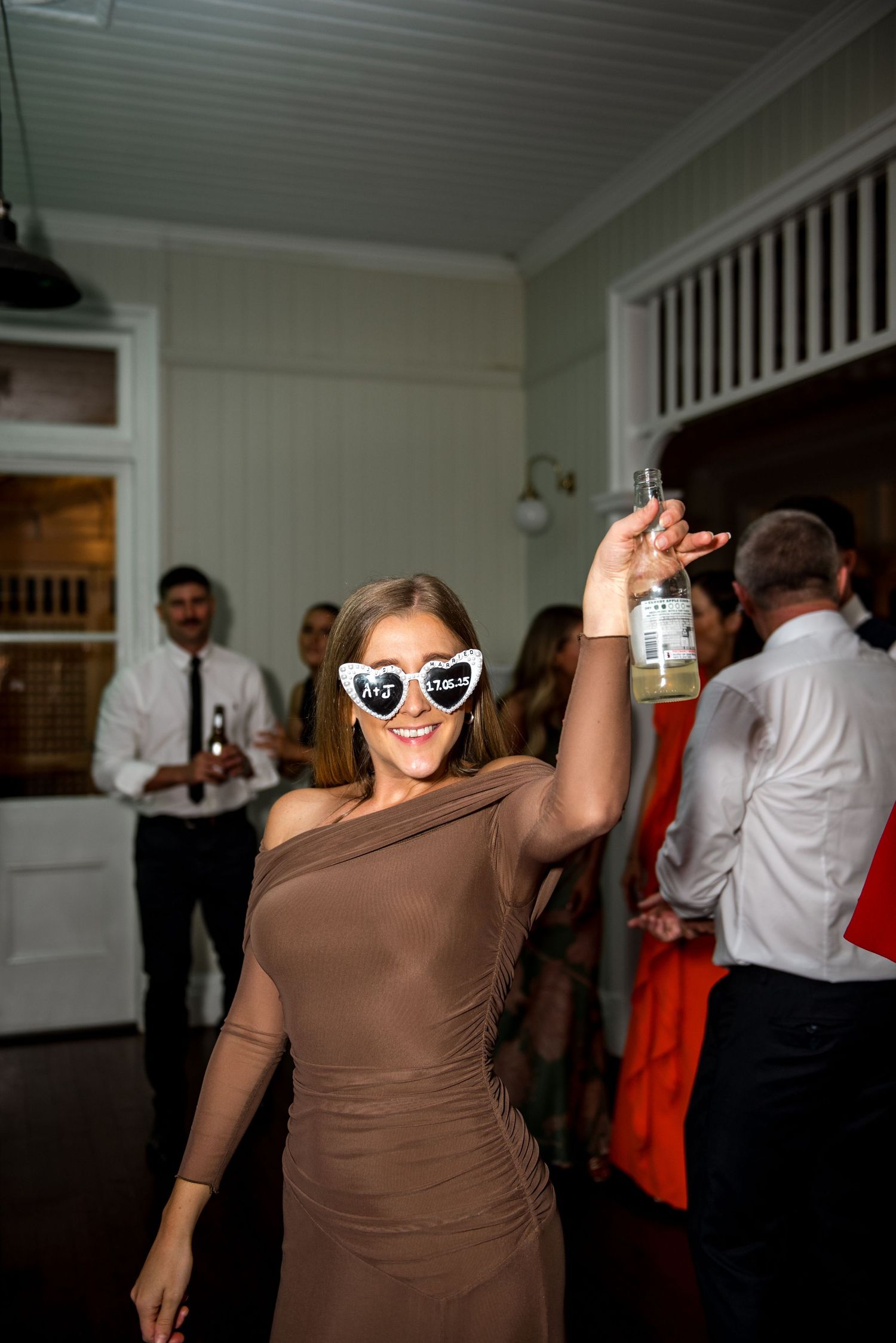 Wedding guest in brown one-shoulder dress wearing novelty sunglasses raising a drink on the dance floor.