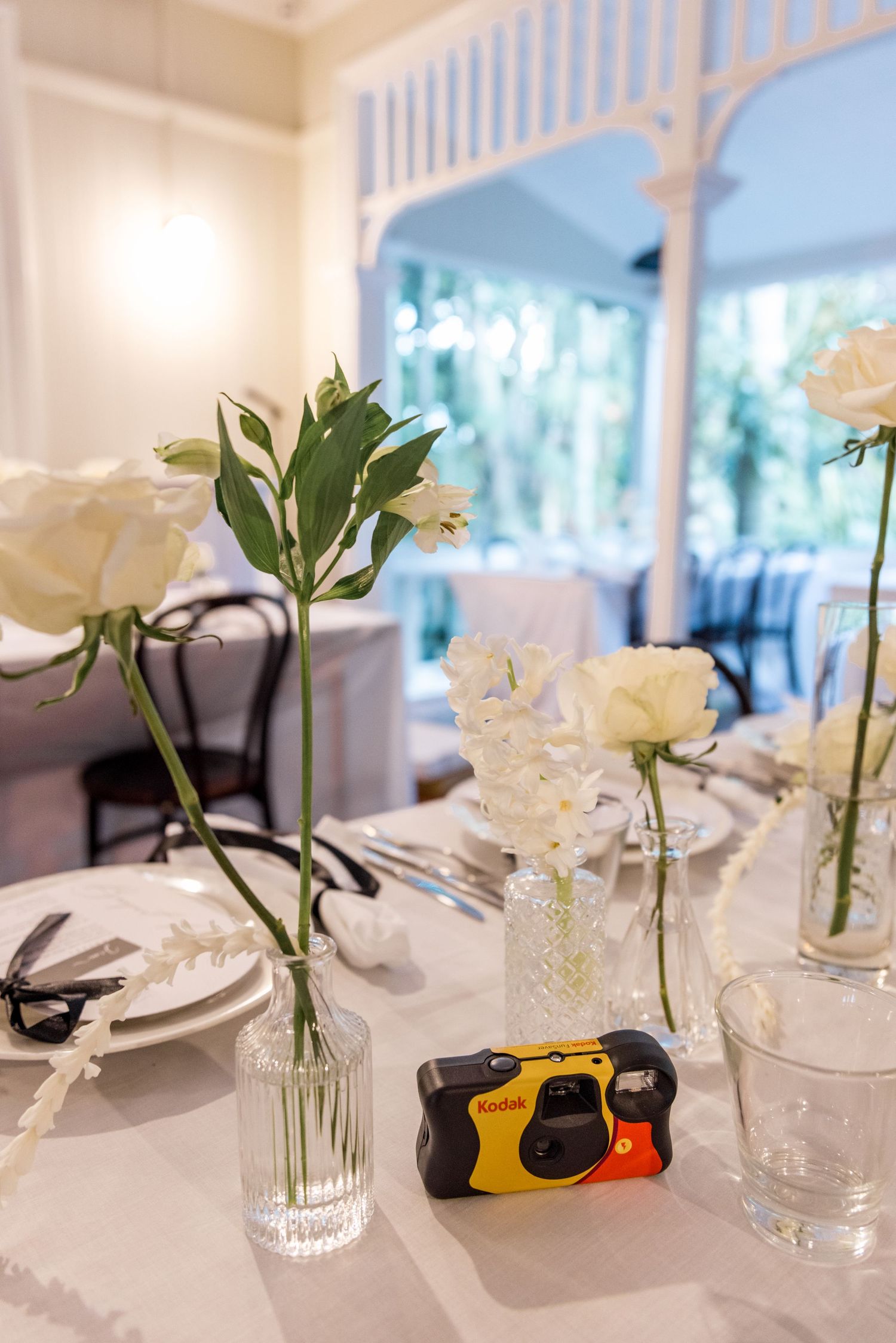Elegant wedding table setting with white roses in glass vases and a Kodak disposable camera as decor.