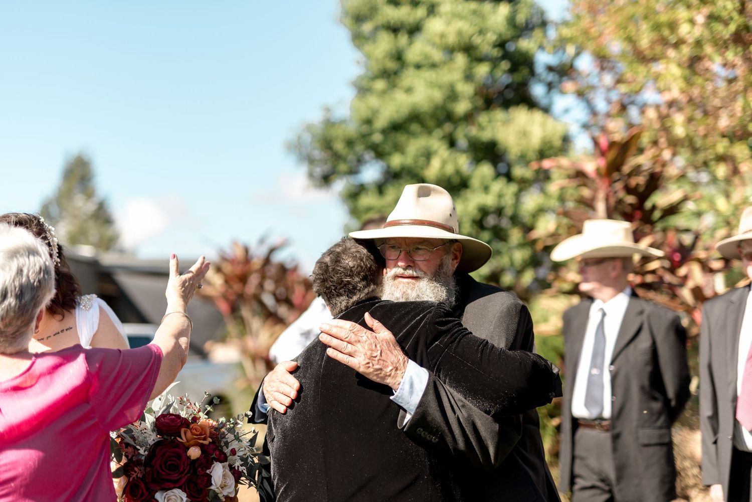 Wedding guest in tan hat shares heartfelt hug with groom in black suit at outdoor ceremony.