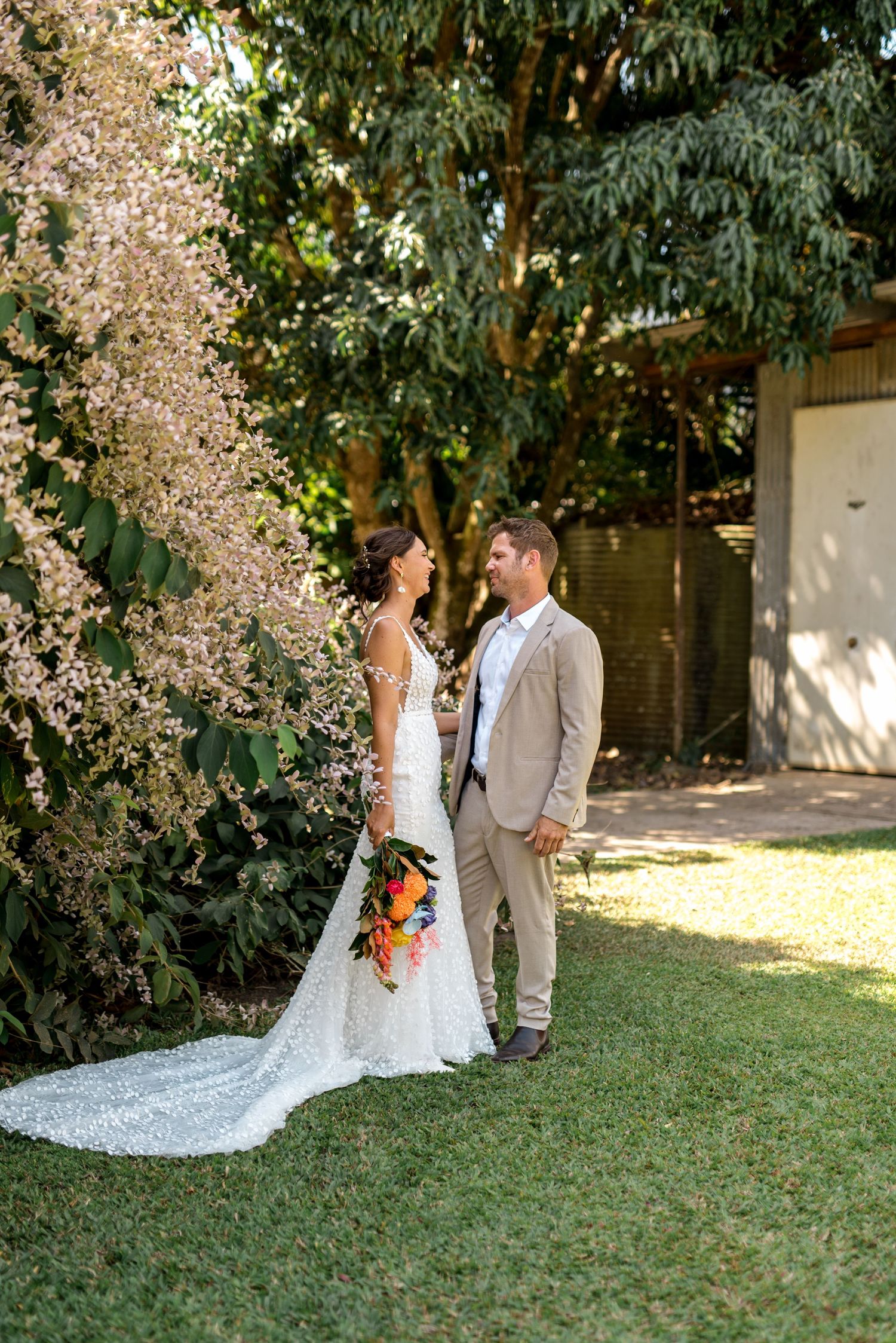 Bride holding vibrant bouquet stands with groom beneath cascading blossoms in garden setting.