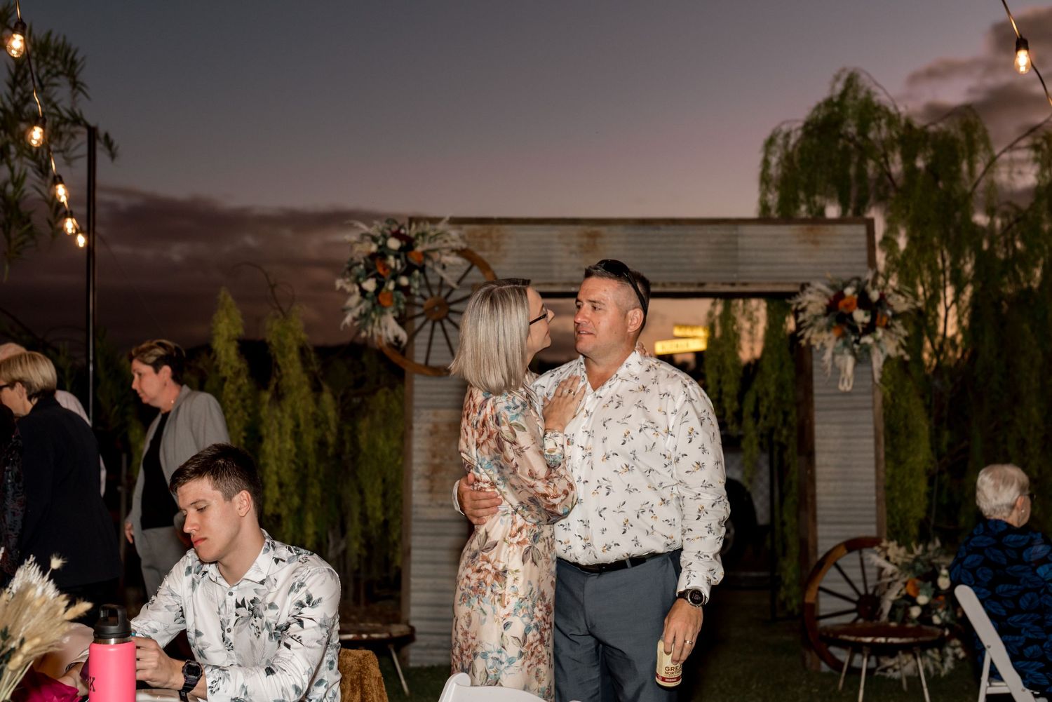 Couple dancing together at an outdoor wedding reception under string lights at dusk.