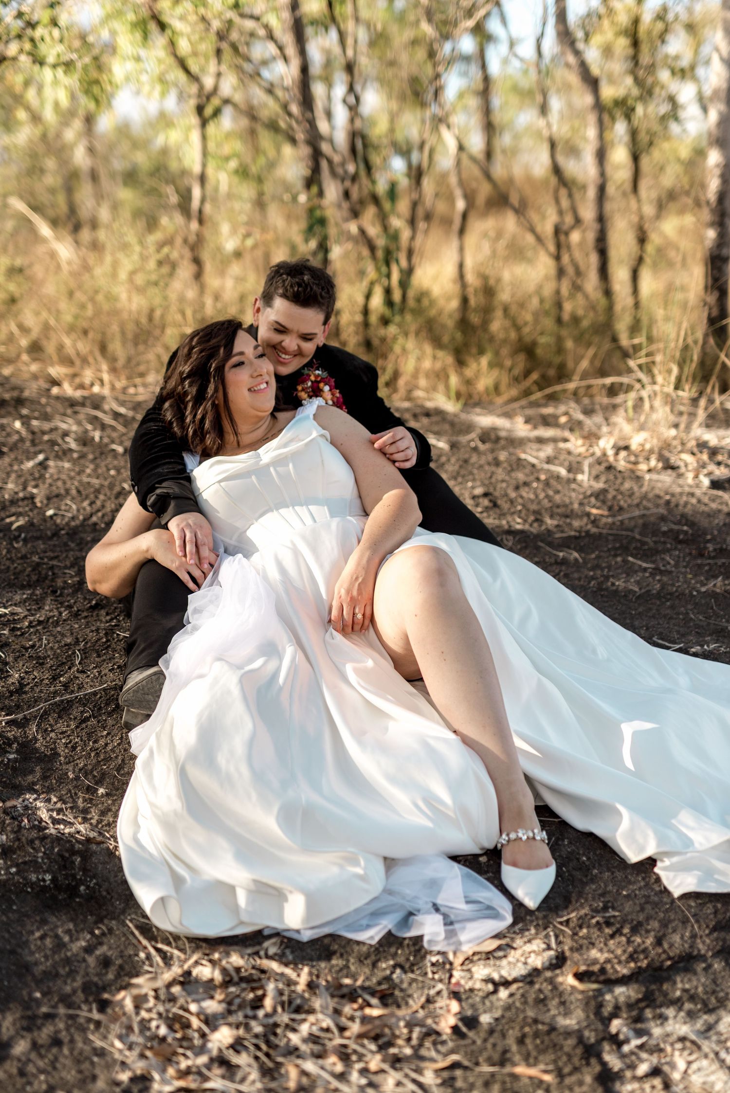 Same-sex couple laughing and relaxing on a rocky outcrop in Australian bush setting, bride in white satin gown.