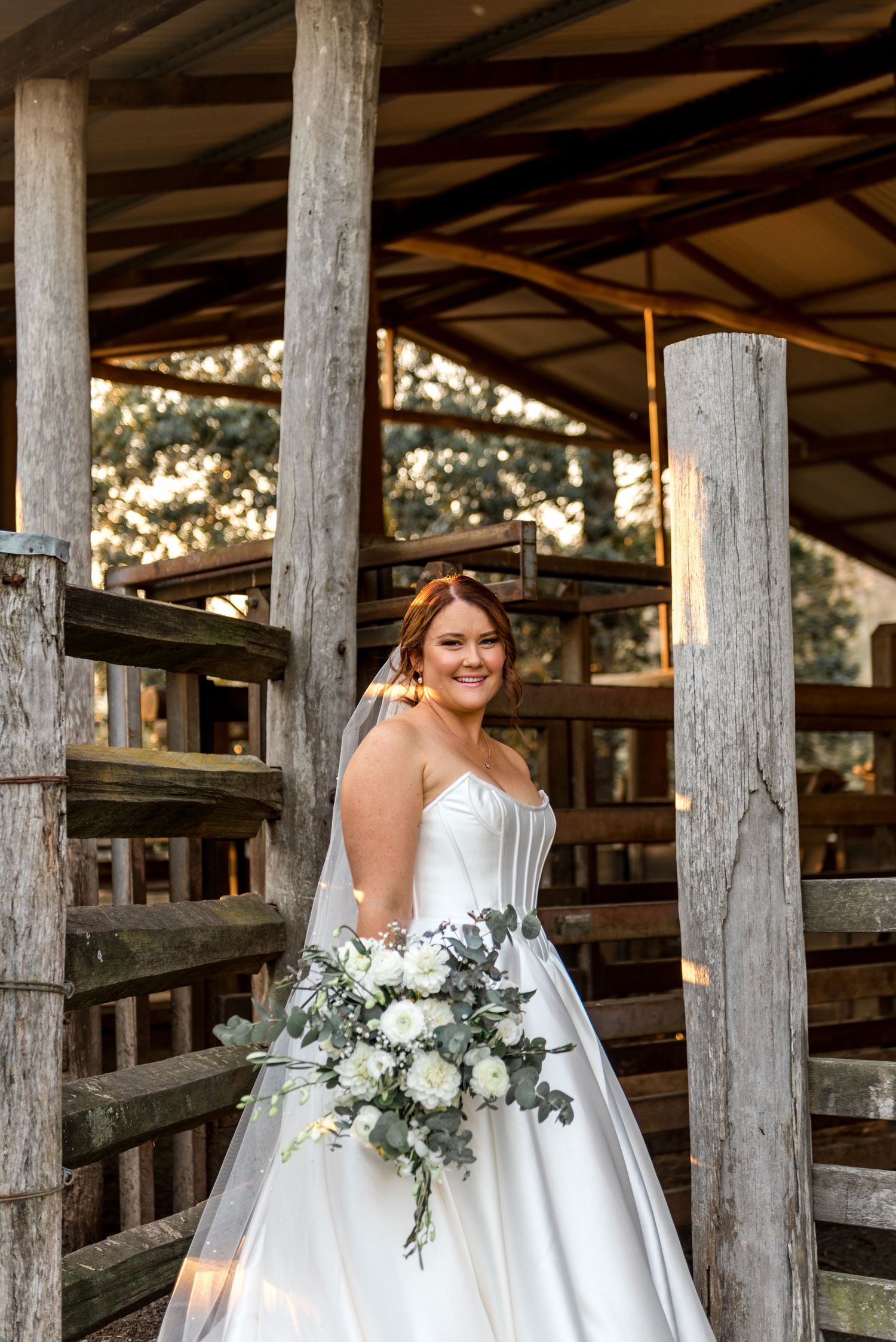 Smiling bride in strapless white ballgown holding white and green bouquet between rustic timber posts.