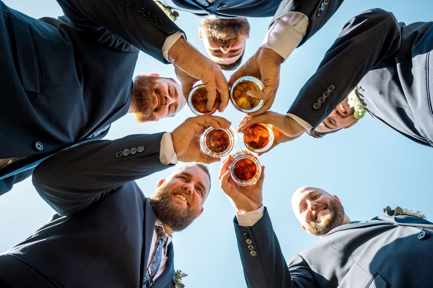 Groomsmen toasting whiskey glasses in circle formation photographed from below against blue sky.