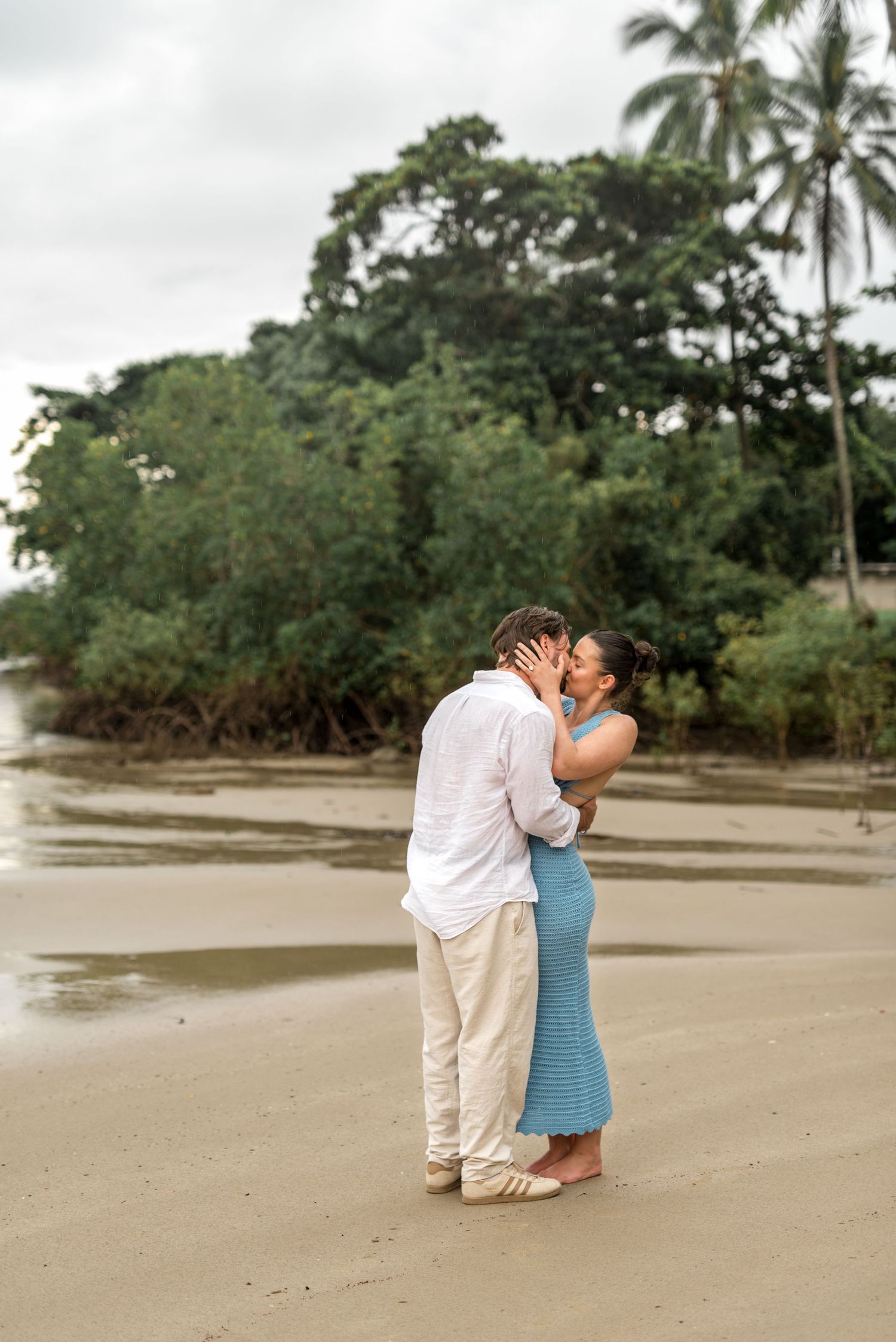 Couple kissing on a sandy beach with dense tropical greenery and palm trees behind them.
