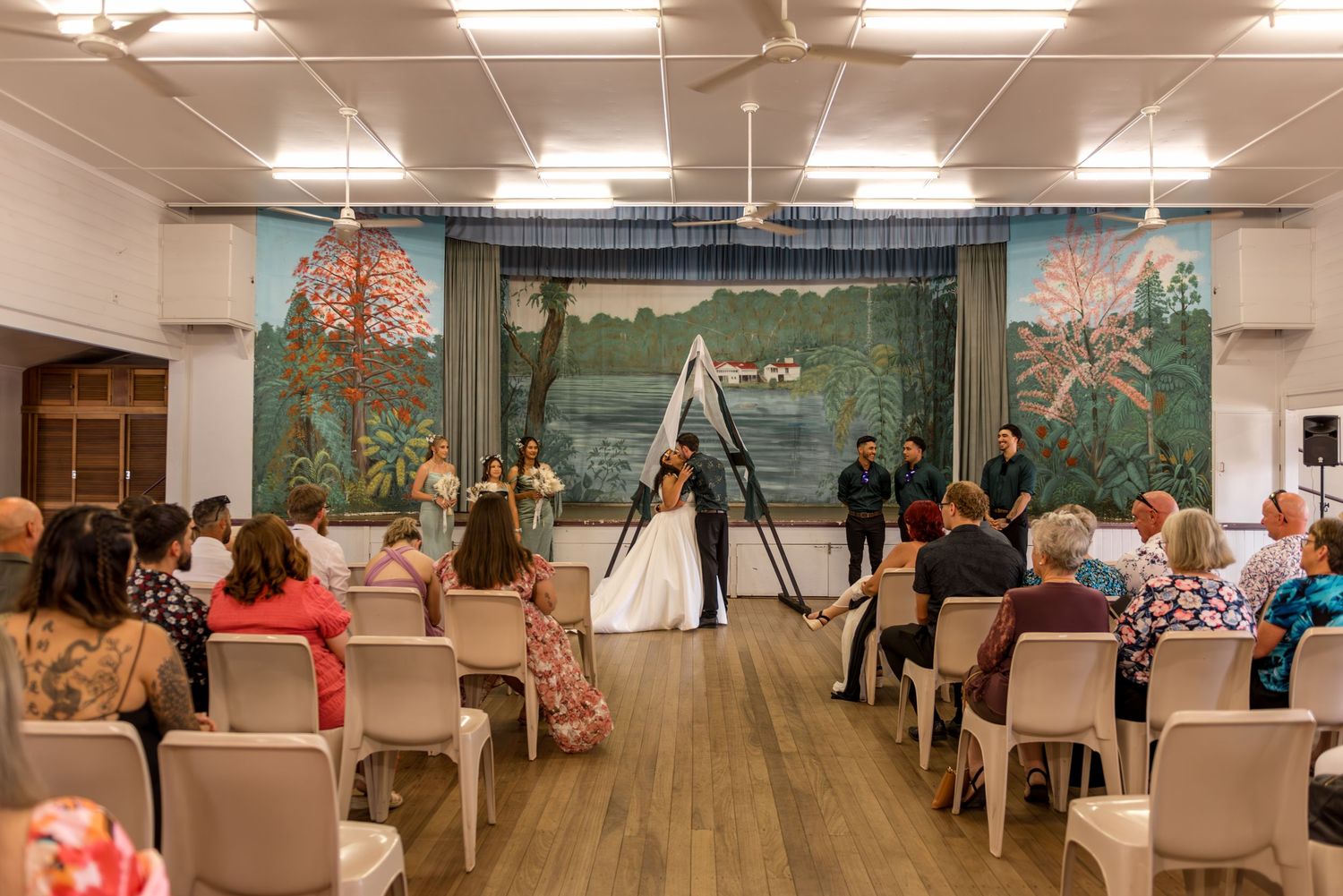 Wide shot of wedding ceremony inside rustic hall with couple at altar, guests seated, and large murals.