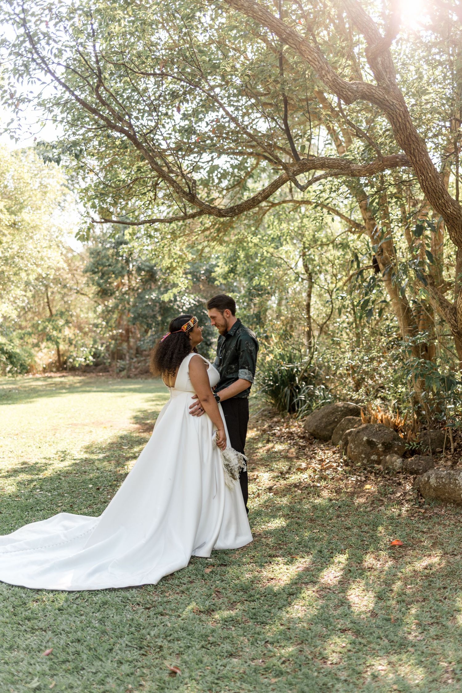 A bride and groom share a romantic moment under a large tree in a sunlit garden.