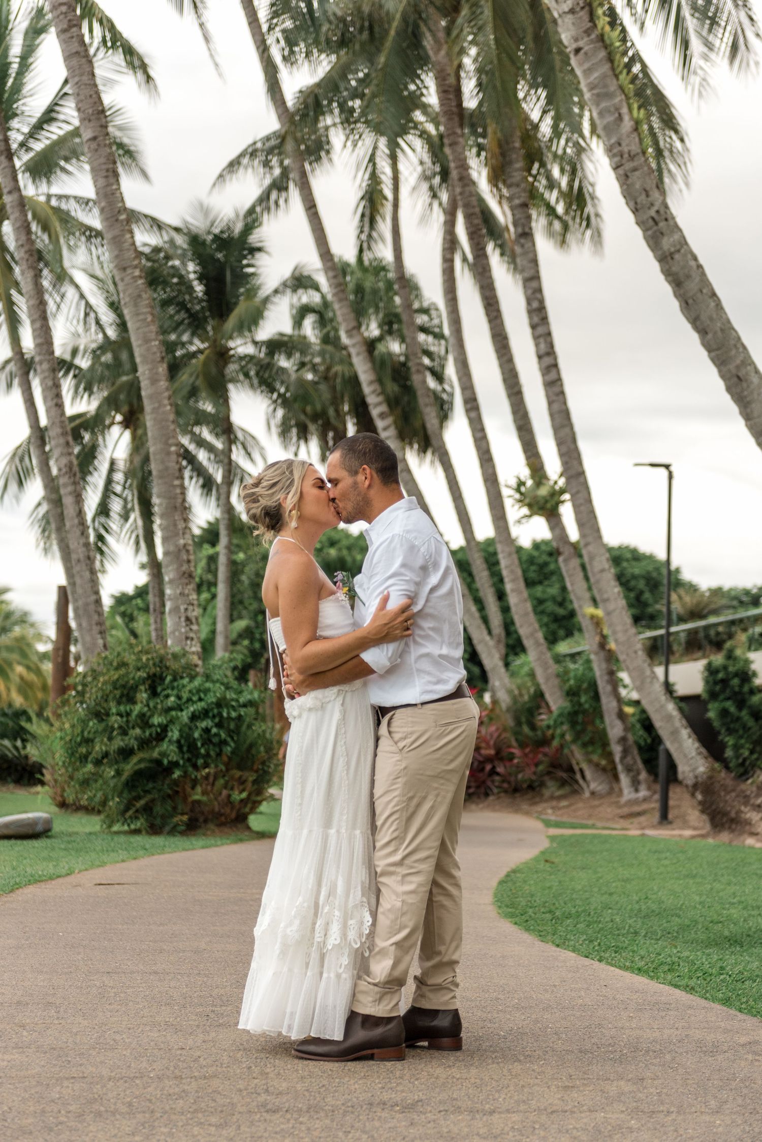 Bride and groom kissing on a curved pathway lined with lush palm trees at a tropical outdoor wedding.