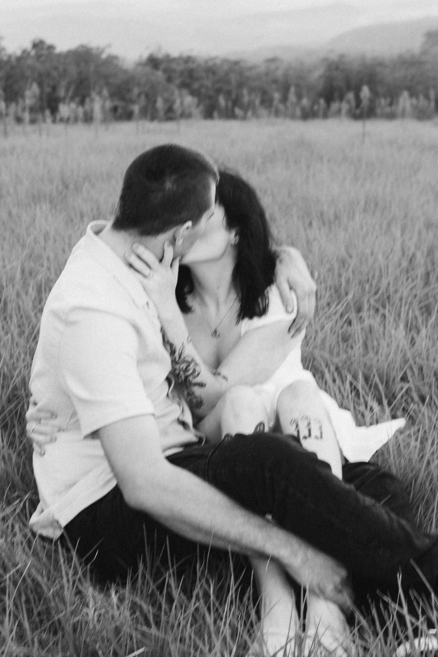 Black and white photo of couple sharing intimate kiss while seated in tall grass field.