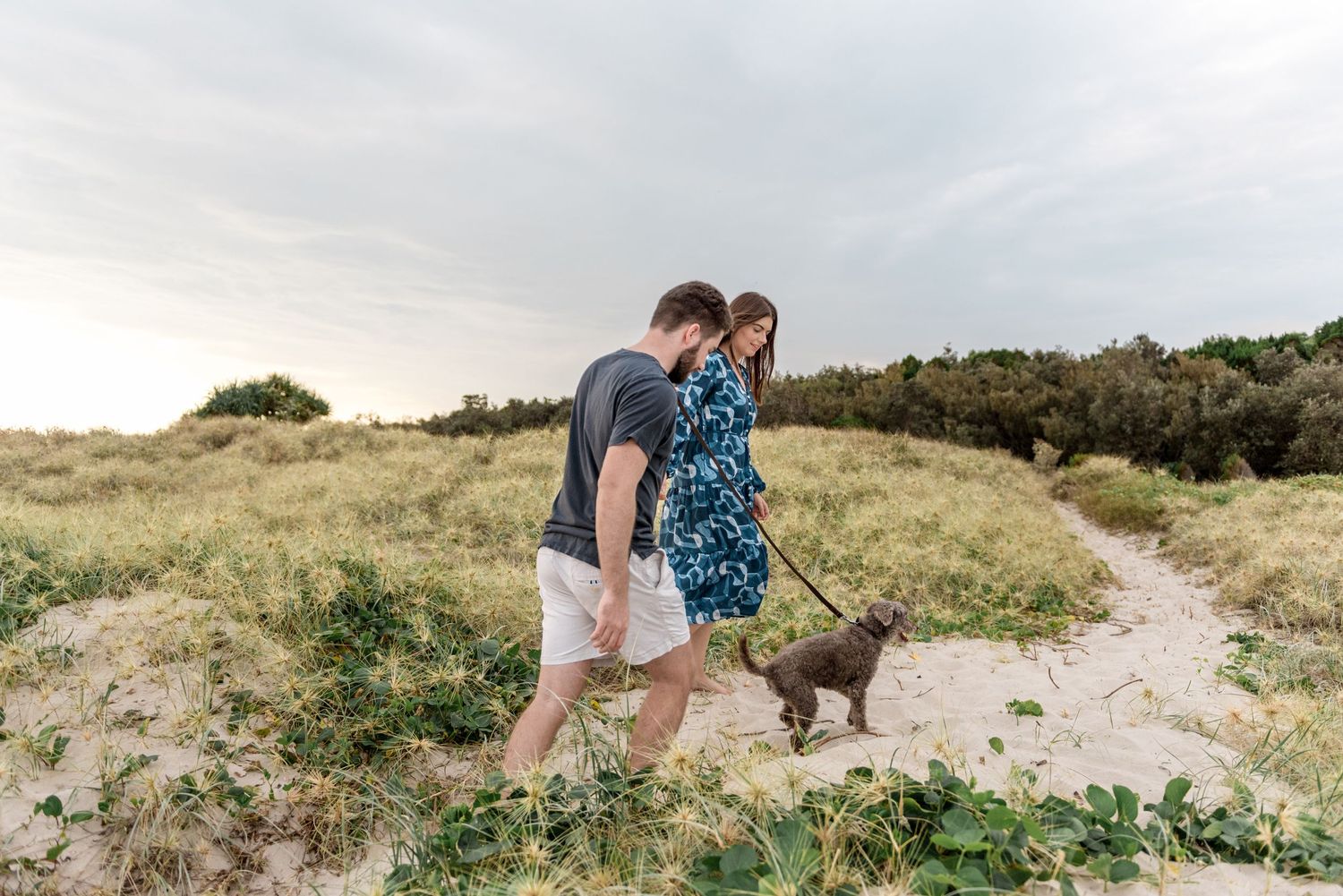 Couple walking grey dog on leash along sandy coastal dune path.