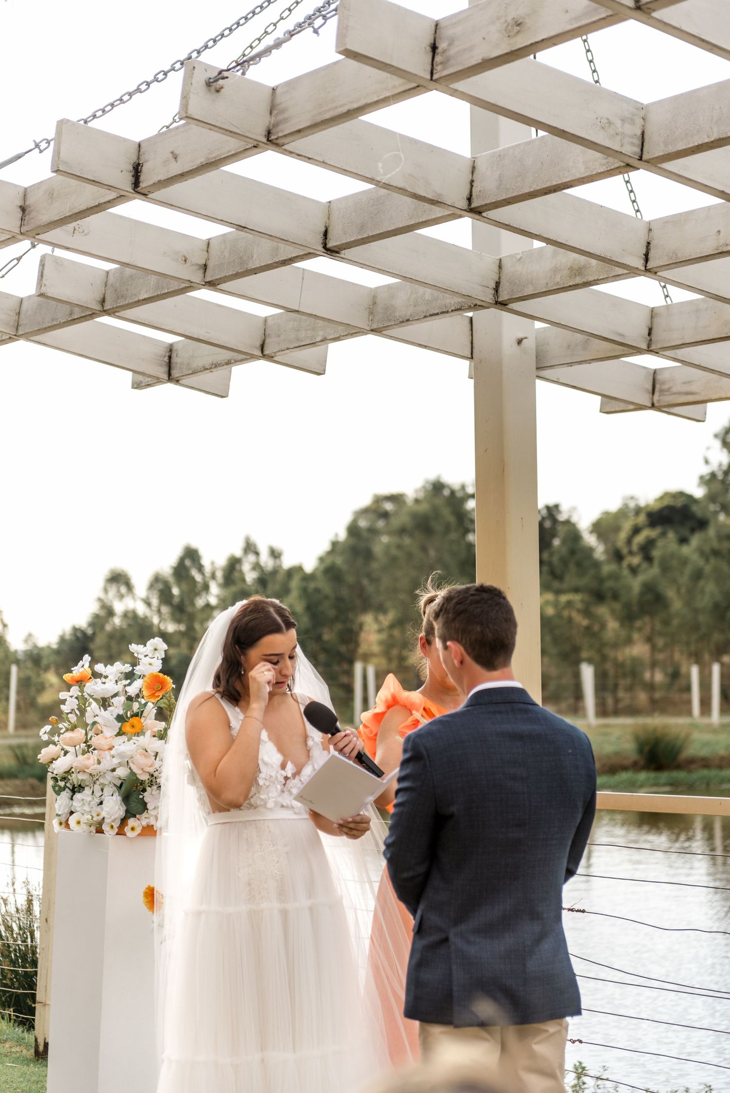 Emotional bride reads vows under a rustic white pergola with orange floral arrangements during outdoor ceremony.