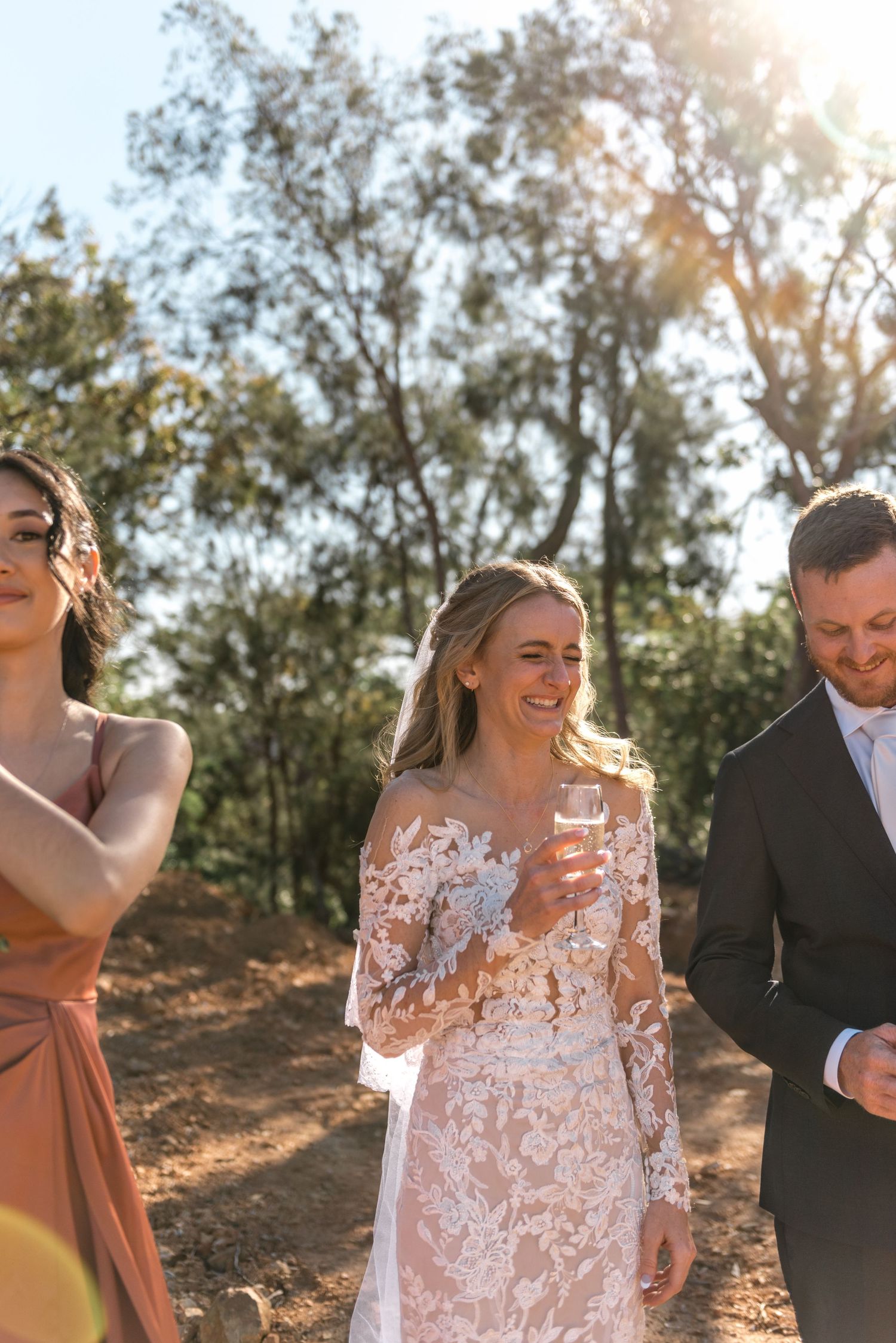 Smiling bride in lace gown holding champagne glass walking with guests in golden sunlight.