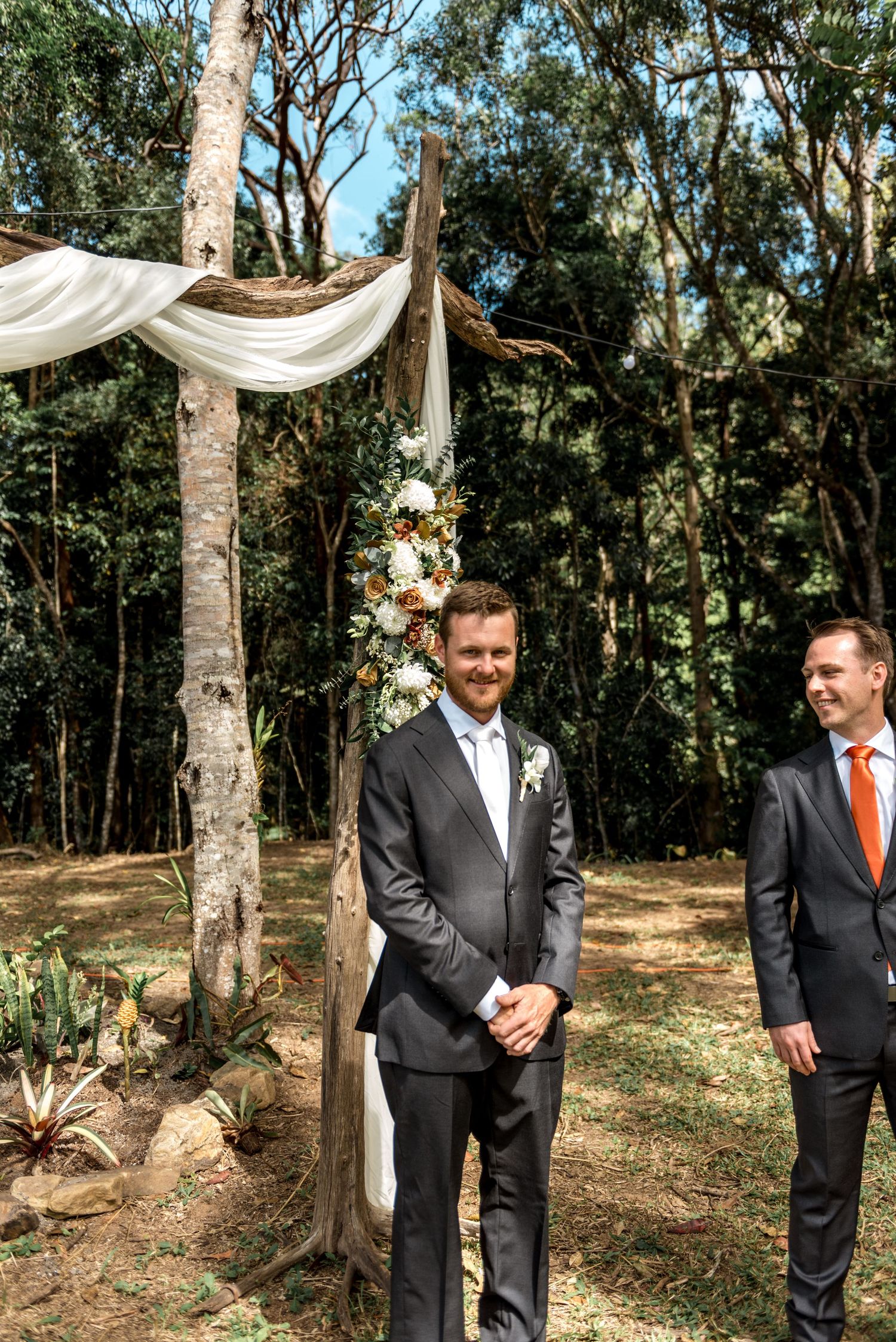 Groom smiling at rustic wooden arch decorated with flowers and white draping in forest setting.