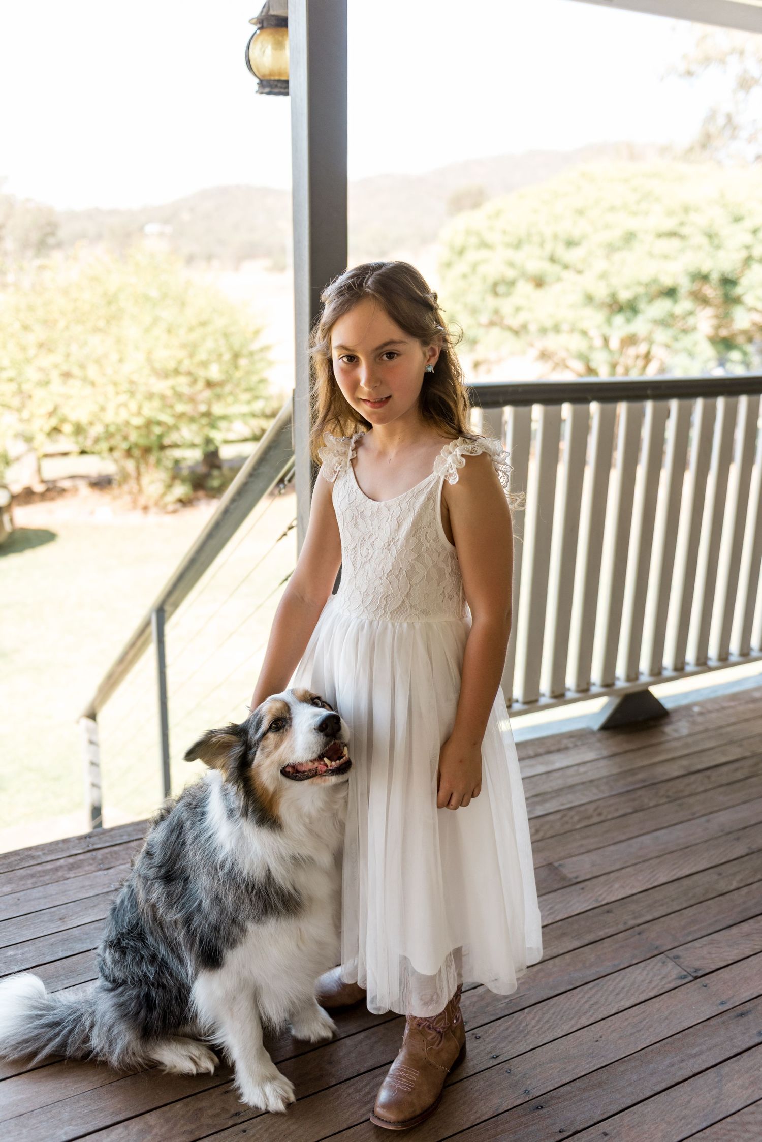 Young flower girl in white lace dress poses with fluffy Australian shepherd dog on rustic porch.