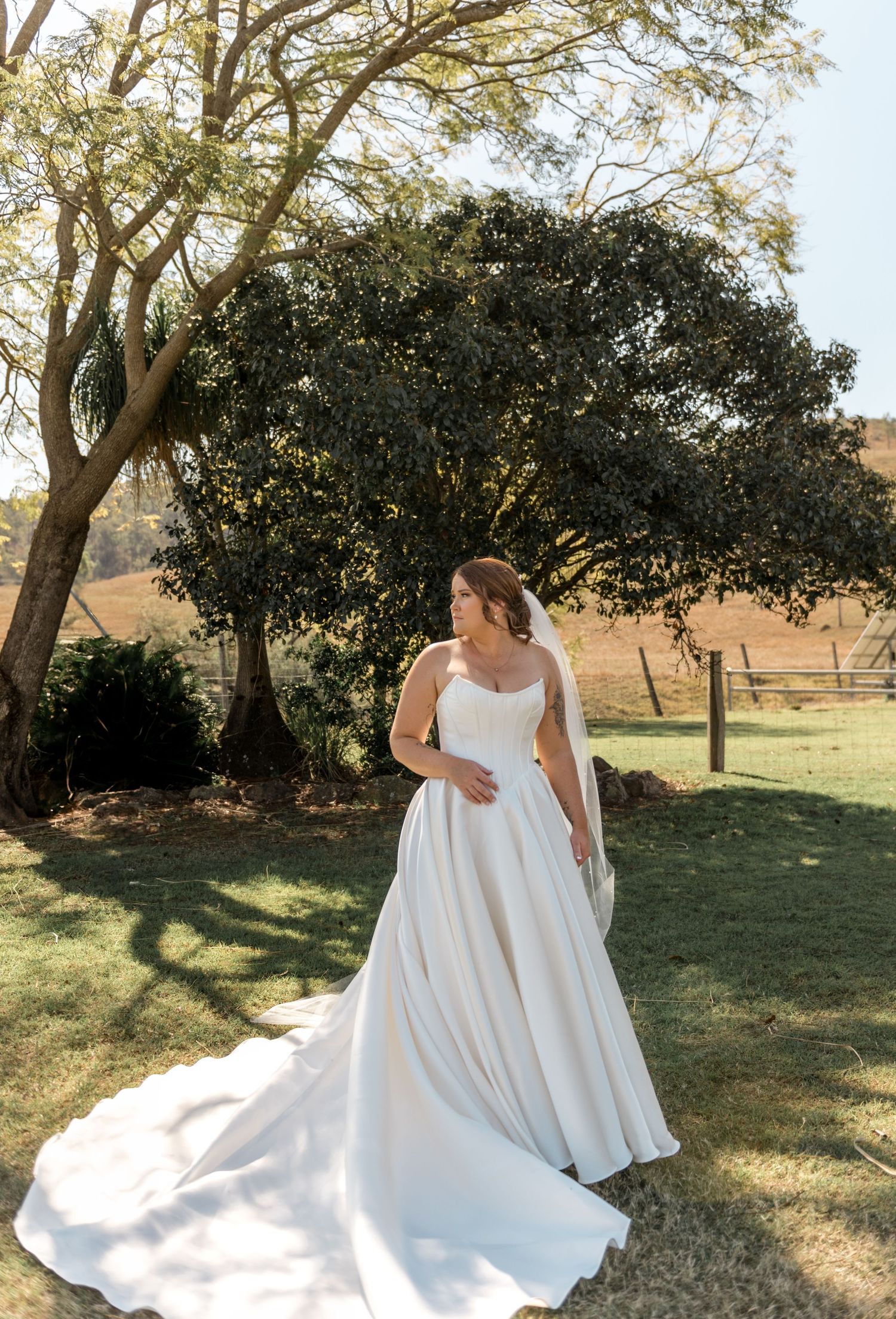 Elegant bride in flowing white satin ballgown with train poses gracefully beneath sunlit garden tree.