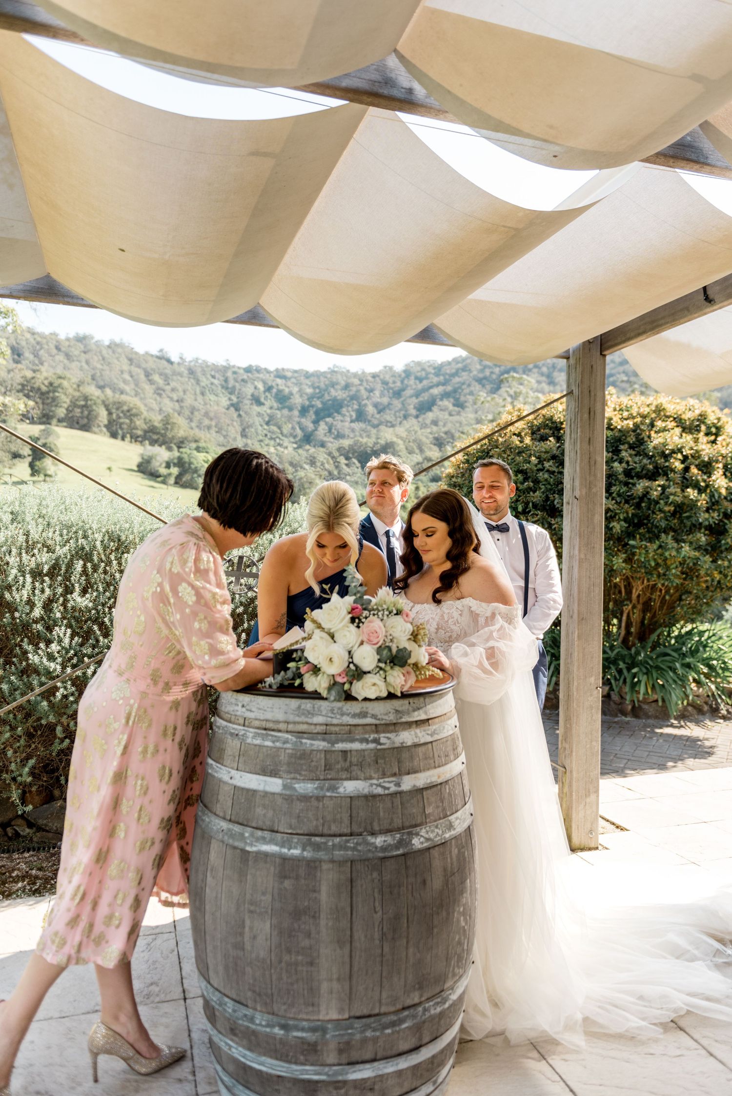 Bride and groom signing marriage documents on rustic wine barrel at outdoor wedding with scenic valley view.