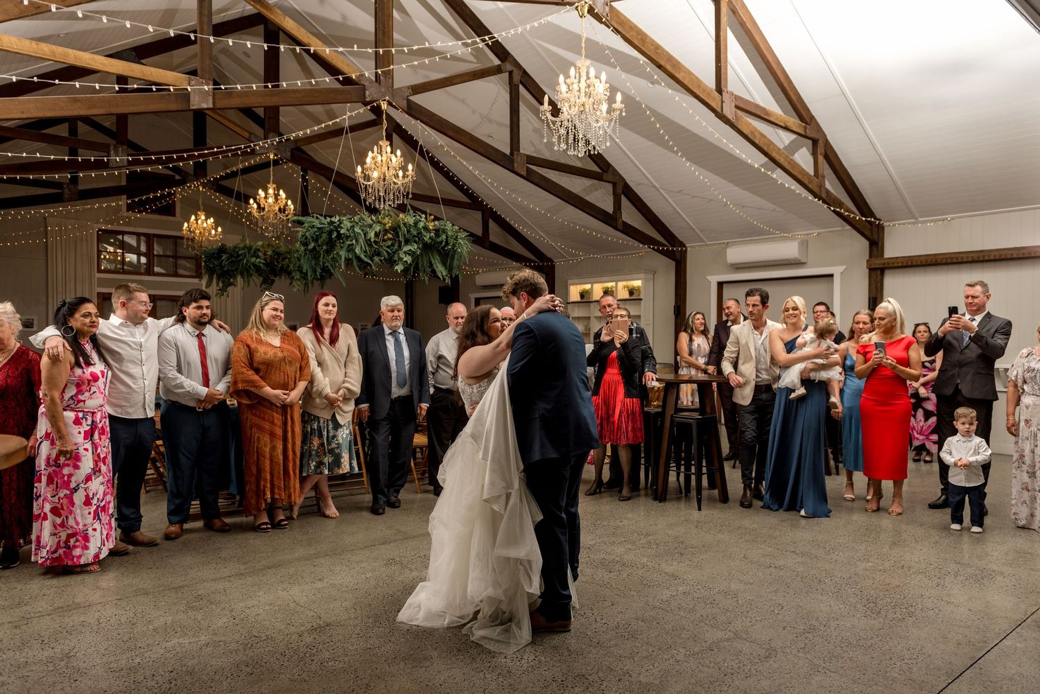 Bride and groom share their first dance in a rustic barn venue surrounded by wedding guests.