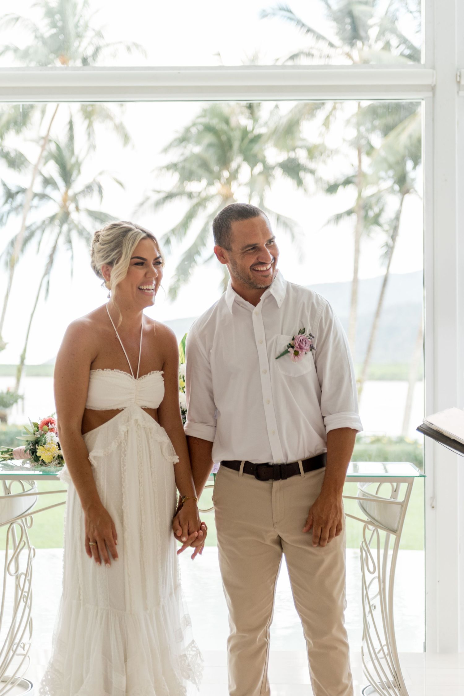 Smiling bride in white halter gown and groom in white shirt holding hands at tropical wedding venue.