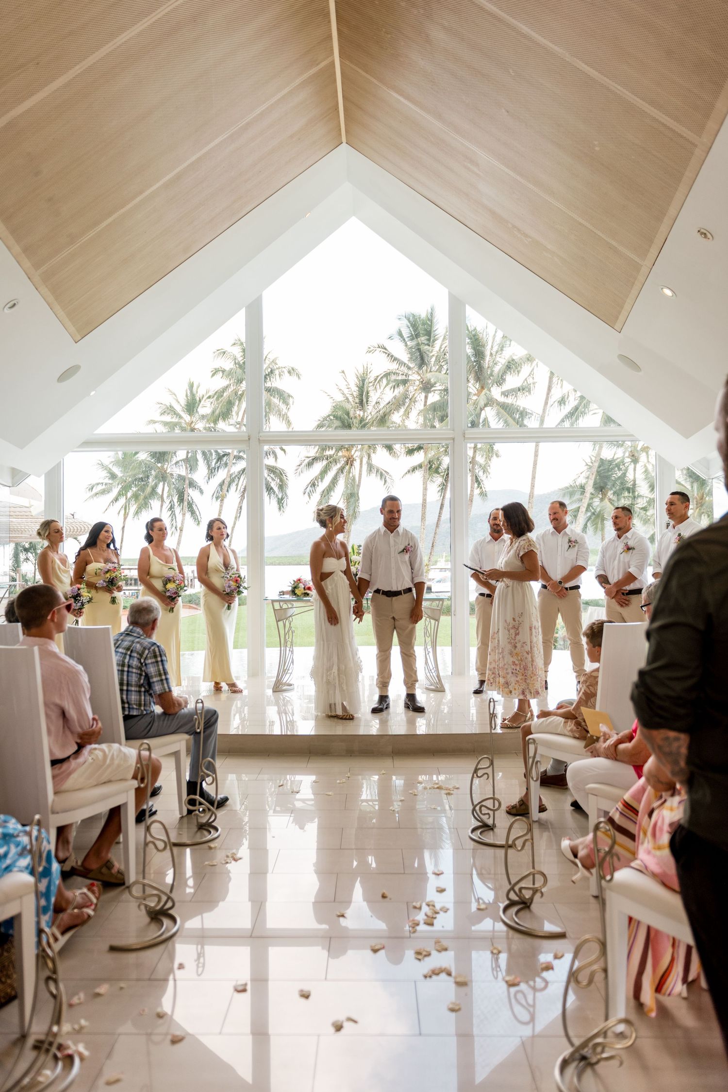 Wide-angle view of a tropical wedding ceremony in a chapel with floor-to-ceiling windows and palm trees.