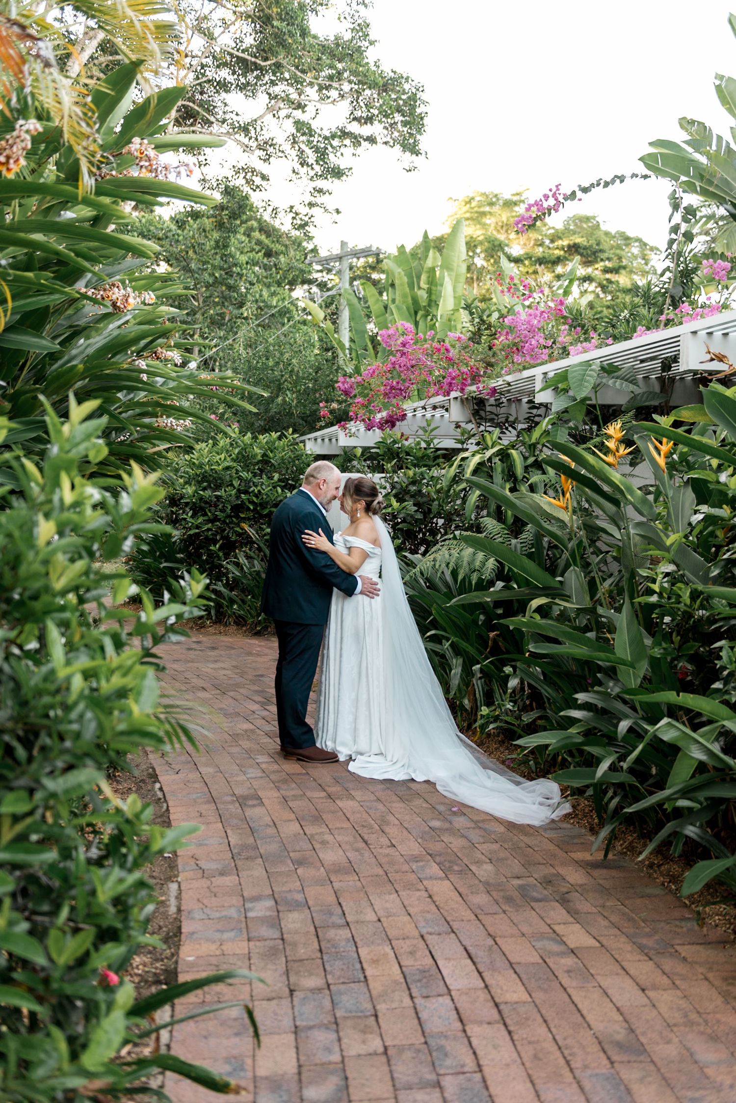 Bride and groom share first look kiss on brick path surrounded by tropical garden blooms.