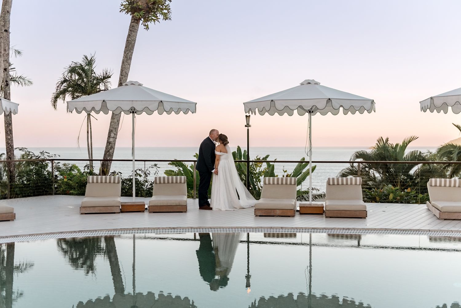 Newlyweds share a romantic kiss poolside at sunset surrounded by white umbrellas and tropical ocean scenery.