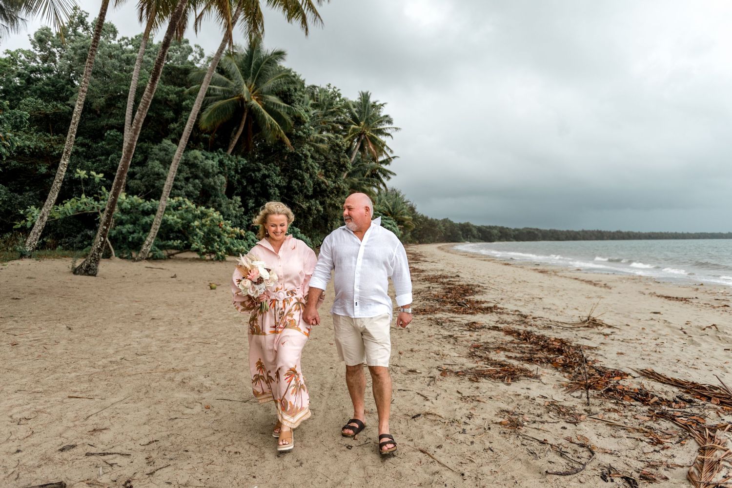 Cheerful mature couple walking together on a sandy tropical beach surrounded by tall palm trees.