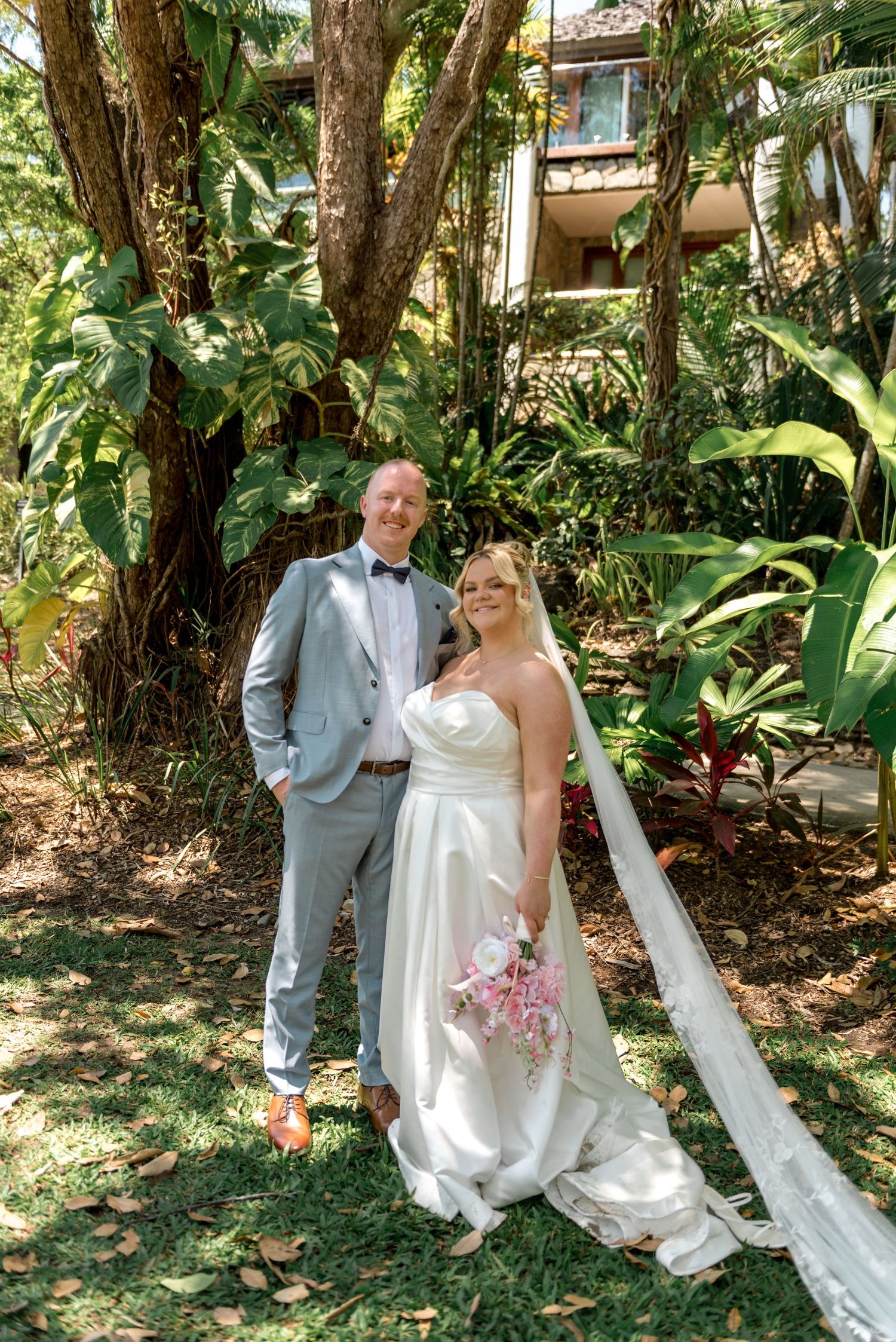 Bride with pink bouquet and groom in grey suit posing in a lush tropical garden setting.