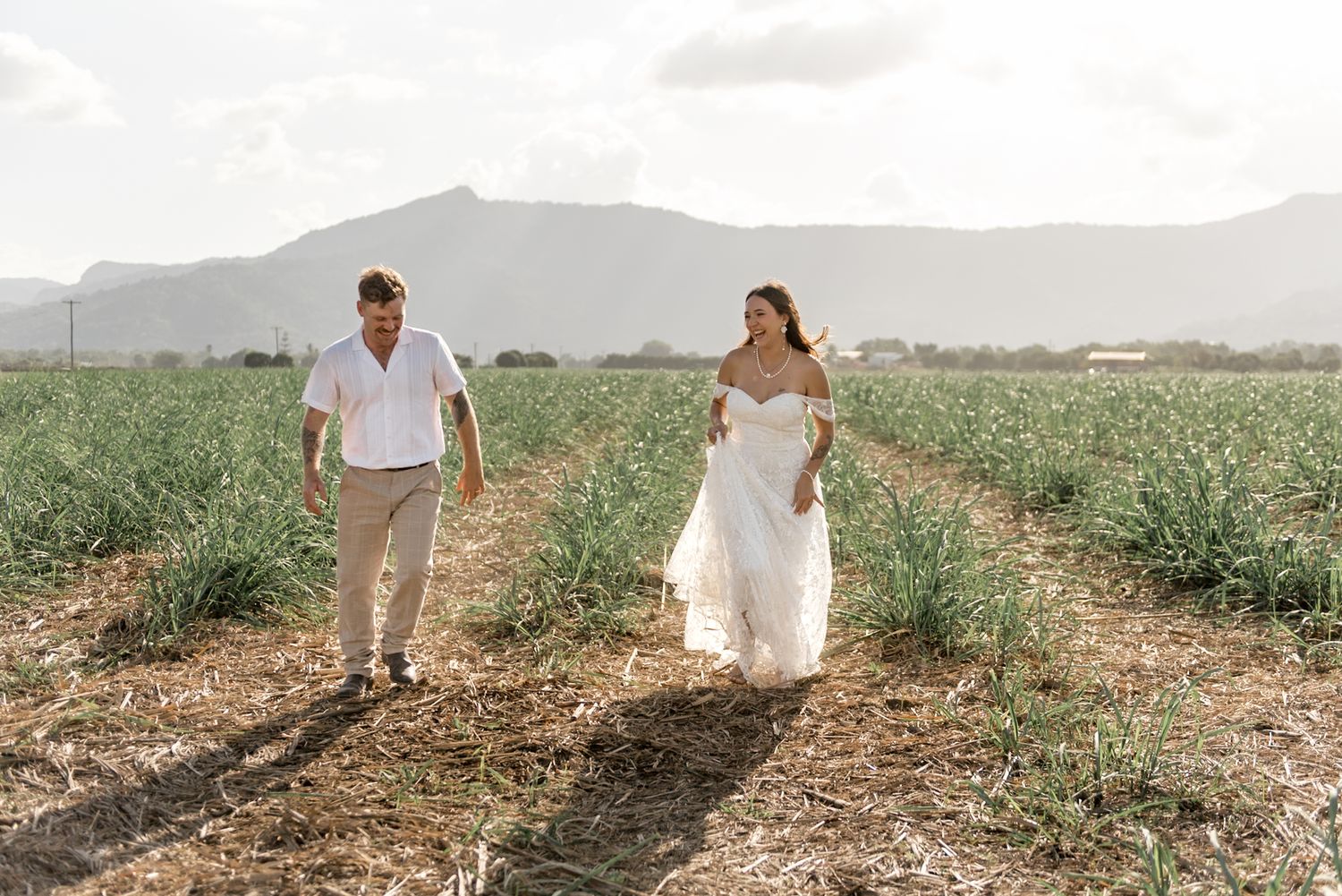 Happy bride and groom walking hand in hand through a green farm field with mountain views.