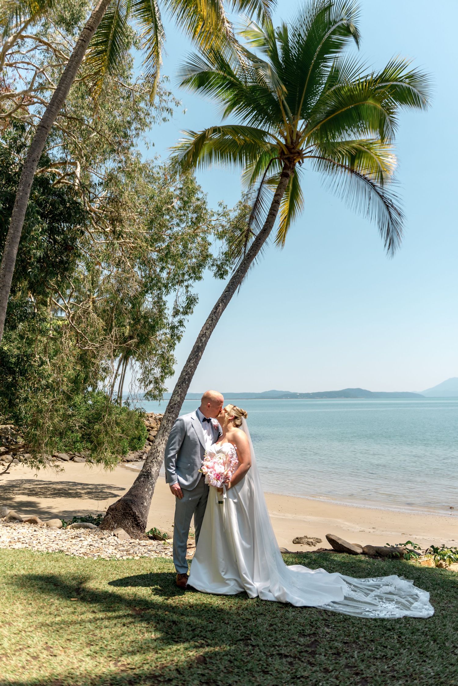 Bride and groom share a romantic kiss under a leaning palm tree on a tropical beach with calm blue ocean views.