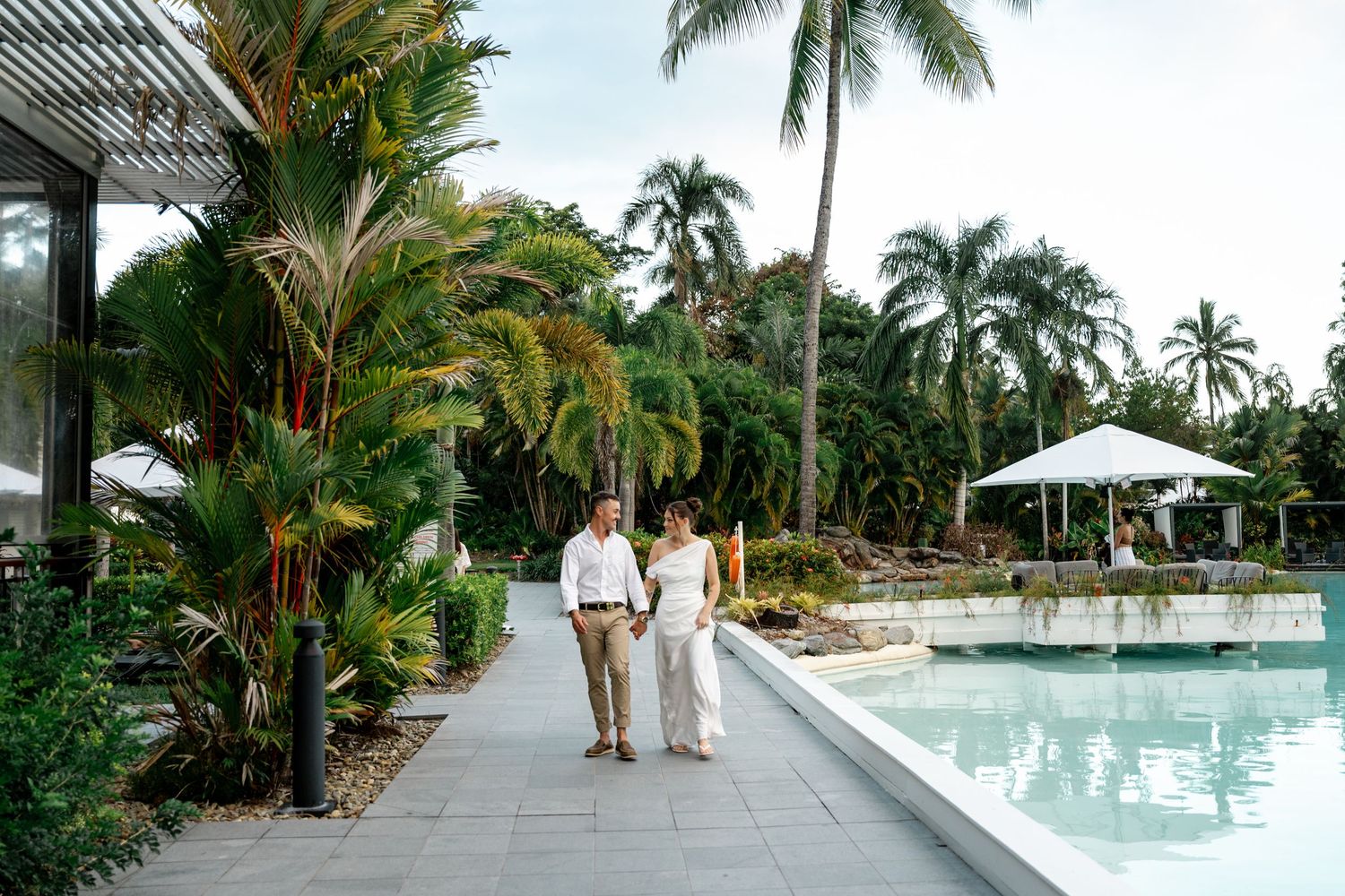 Newlywed couple walking hand in hand along a tropical resort poolside pathway lined with palms at Sheraton Grand Mirage Port Douglas.