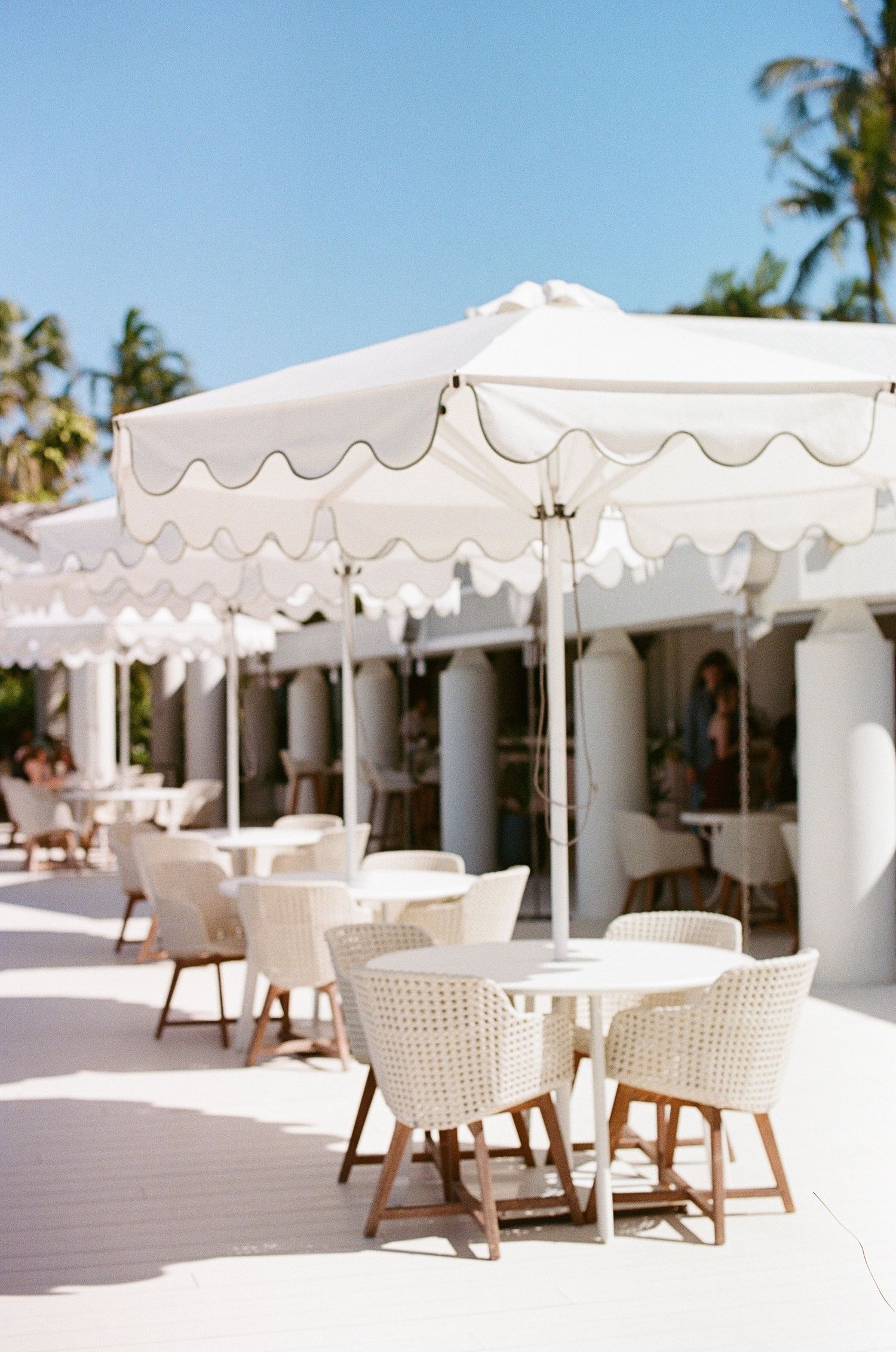 Elegant outdoor dining area with white scalloped umbrellas, wicker chairs, and round tables at a tropical resort.