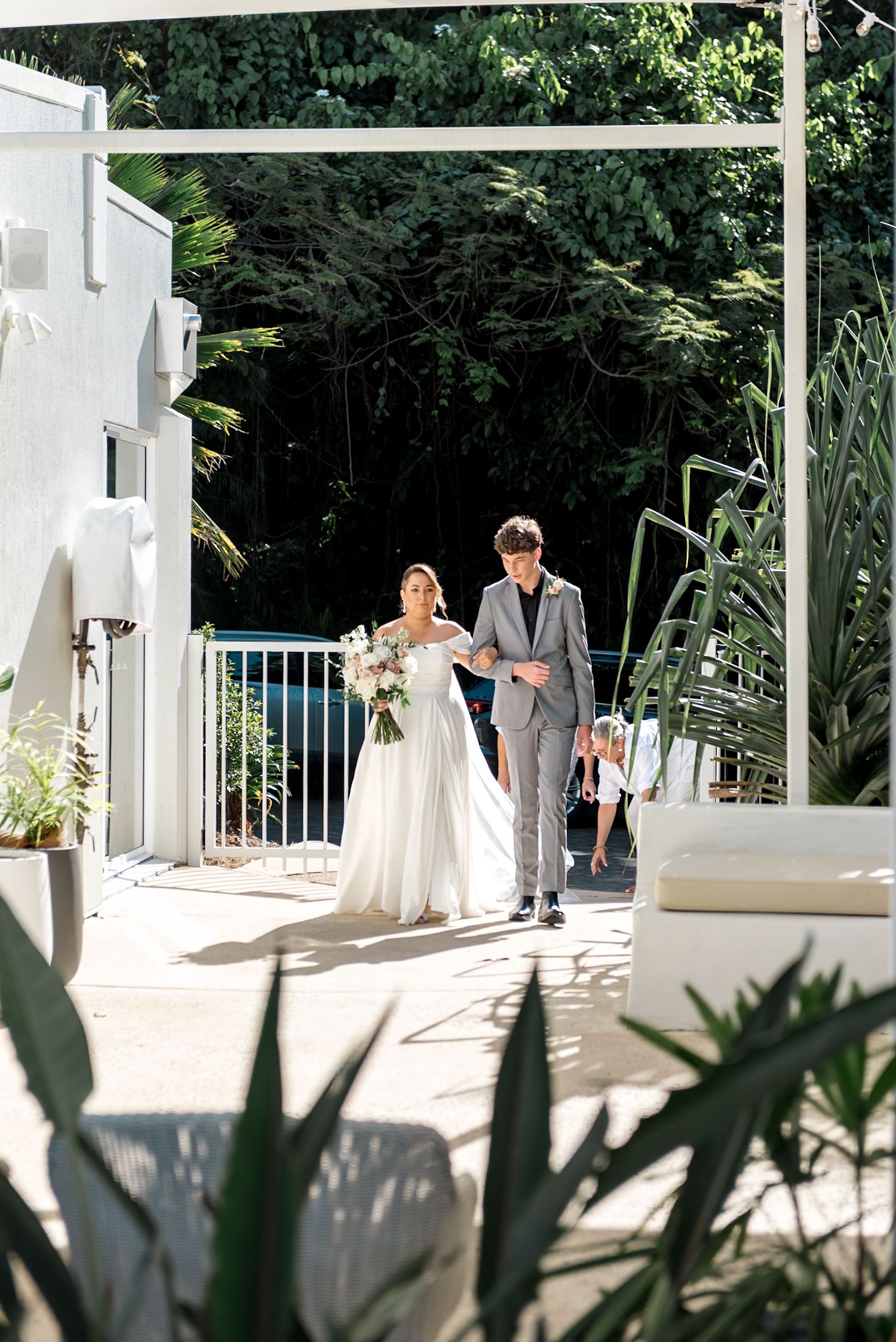 Bride in white gown walks with escort toward tropical outdoor wedding ceremony past pool area.