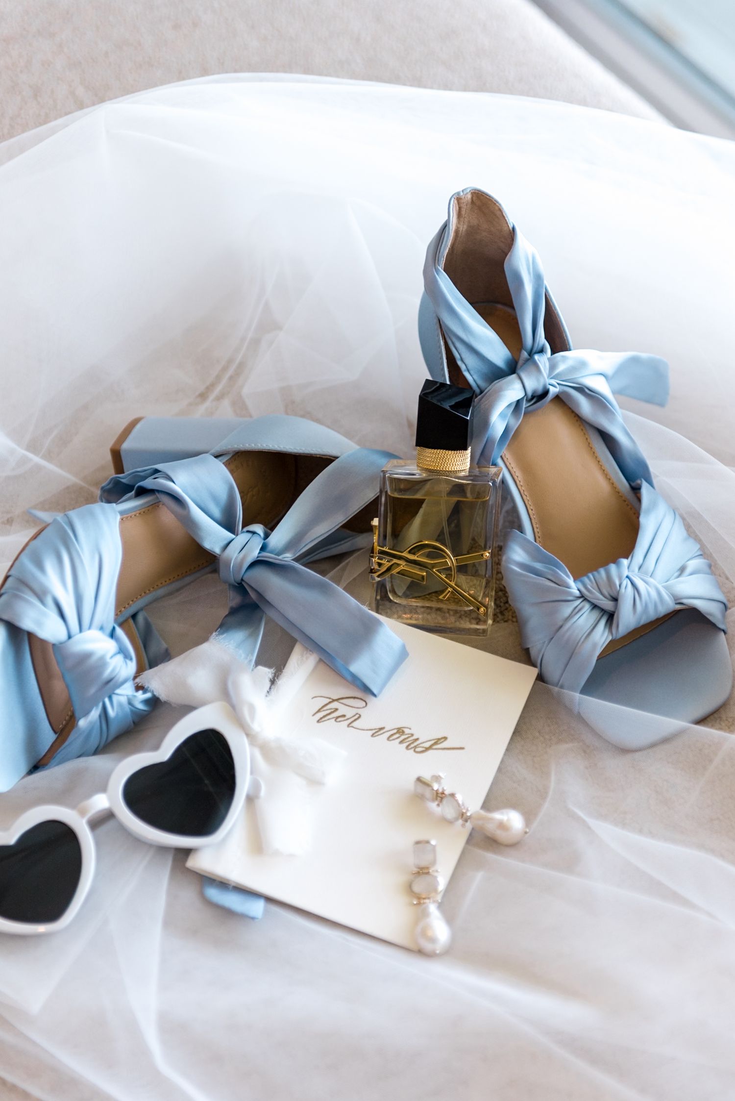Light blue satin bow heels with pearl earrings, YSL perfume, sunglasses, and wedding card on white tulle.