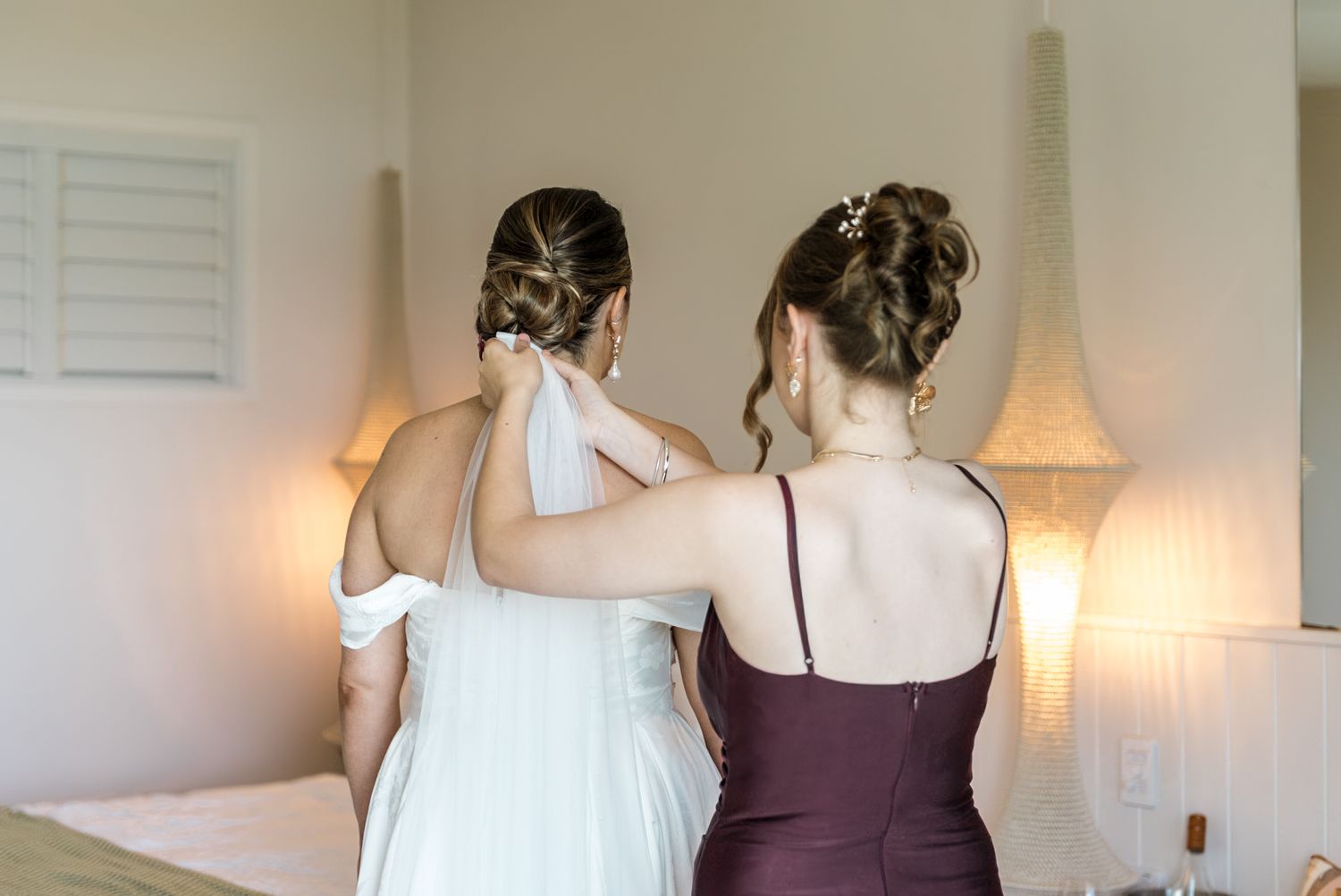 Bridesmaid in burgundy dress adjusting the veil of a bride with an elegant updo from behind.