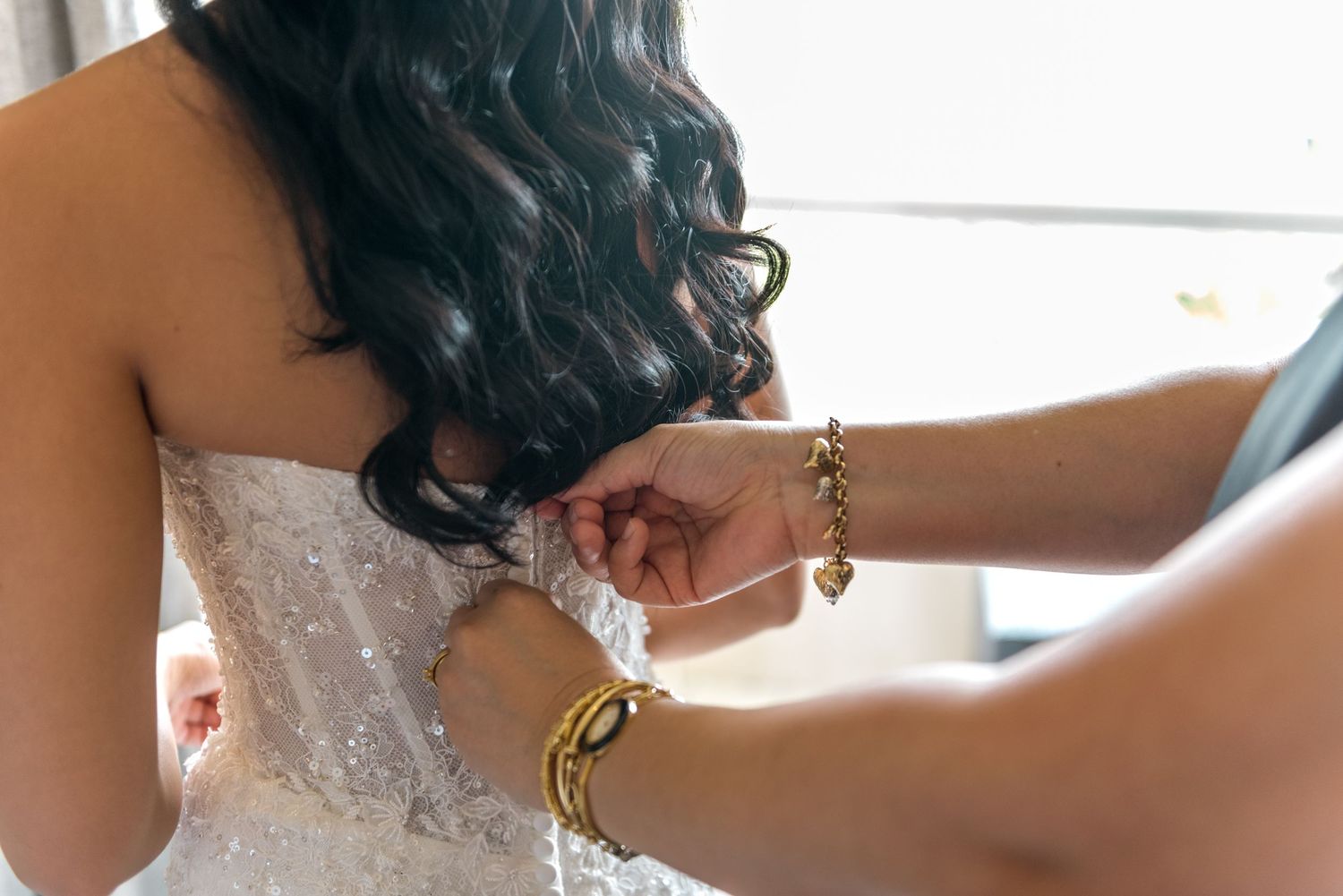 Close-up of hands with gold bracelets fastening buttons on a white lace wedding dress.