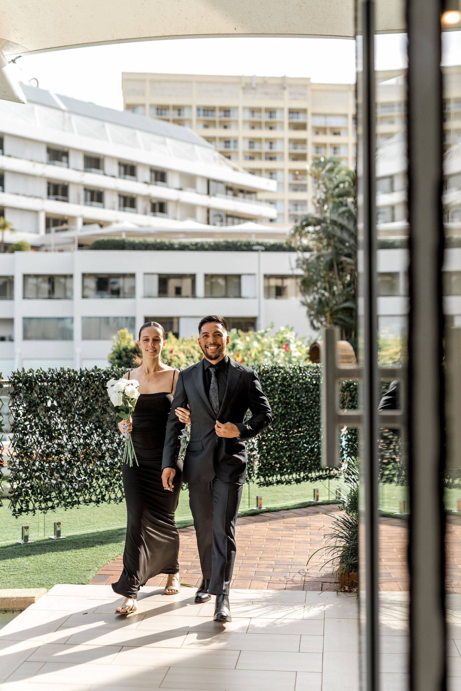 Couple in black formal wear walk together through outdoor hotel wedding venue.