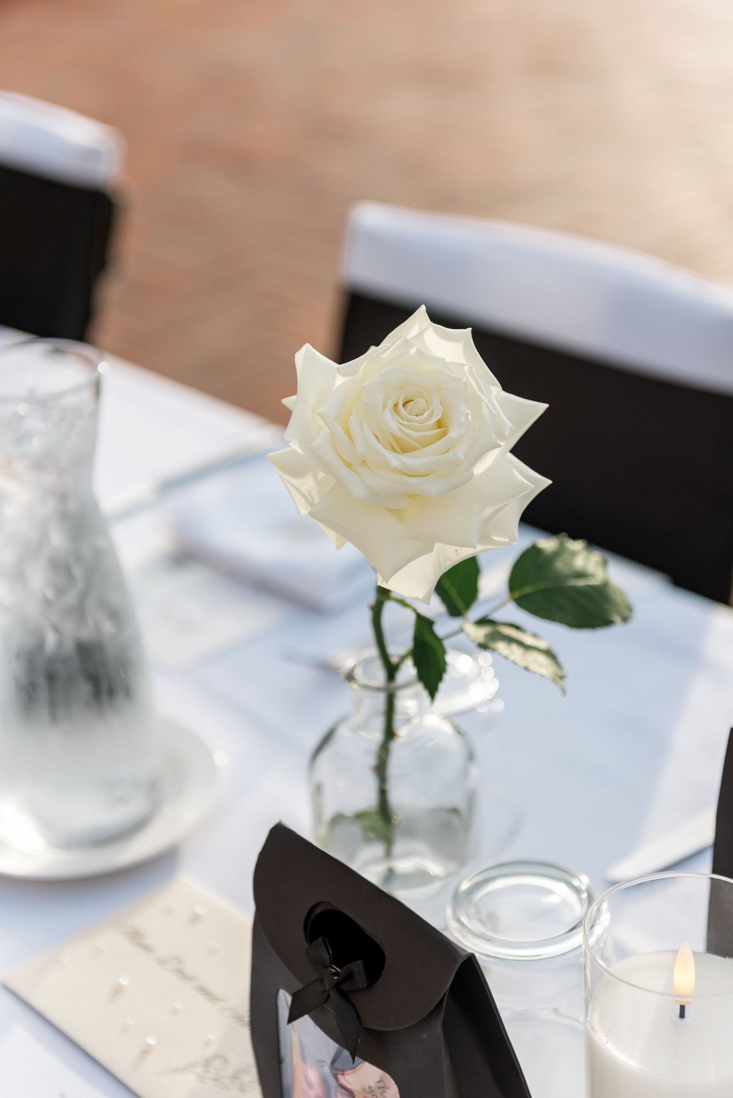 Single white rose in glass vase on white tablecloth wedding reception table with candle.