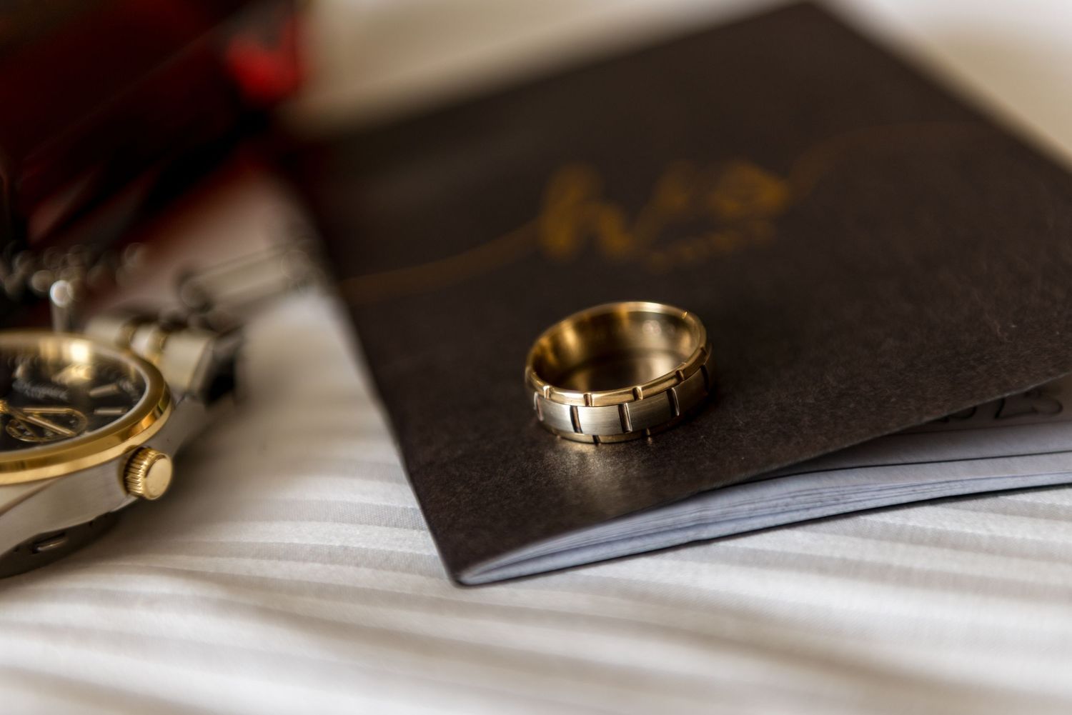 Close-up of a gold wedding band resting on a dark groom's vow book beside a watch on white linen.