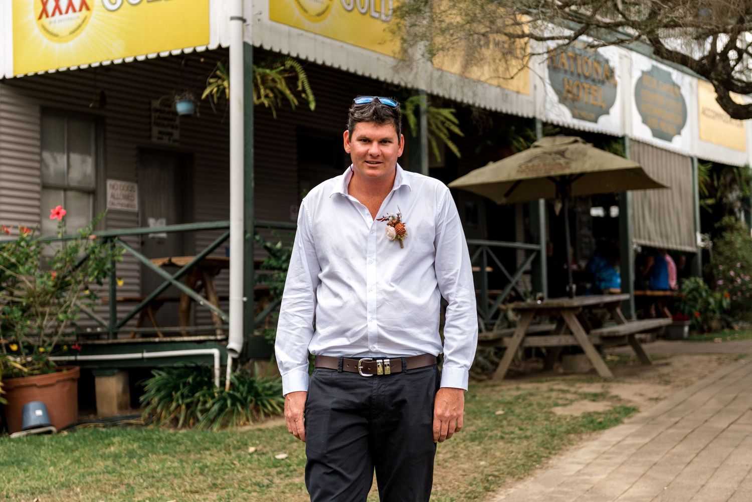 Groom in white shirt with boutonniere standing outside a country pub with XXXX Gold signage.