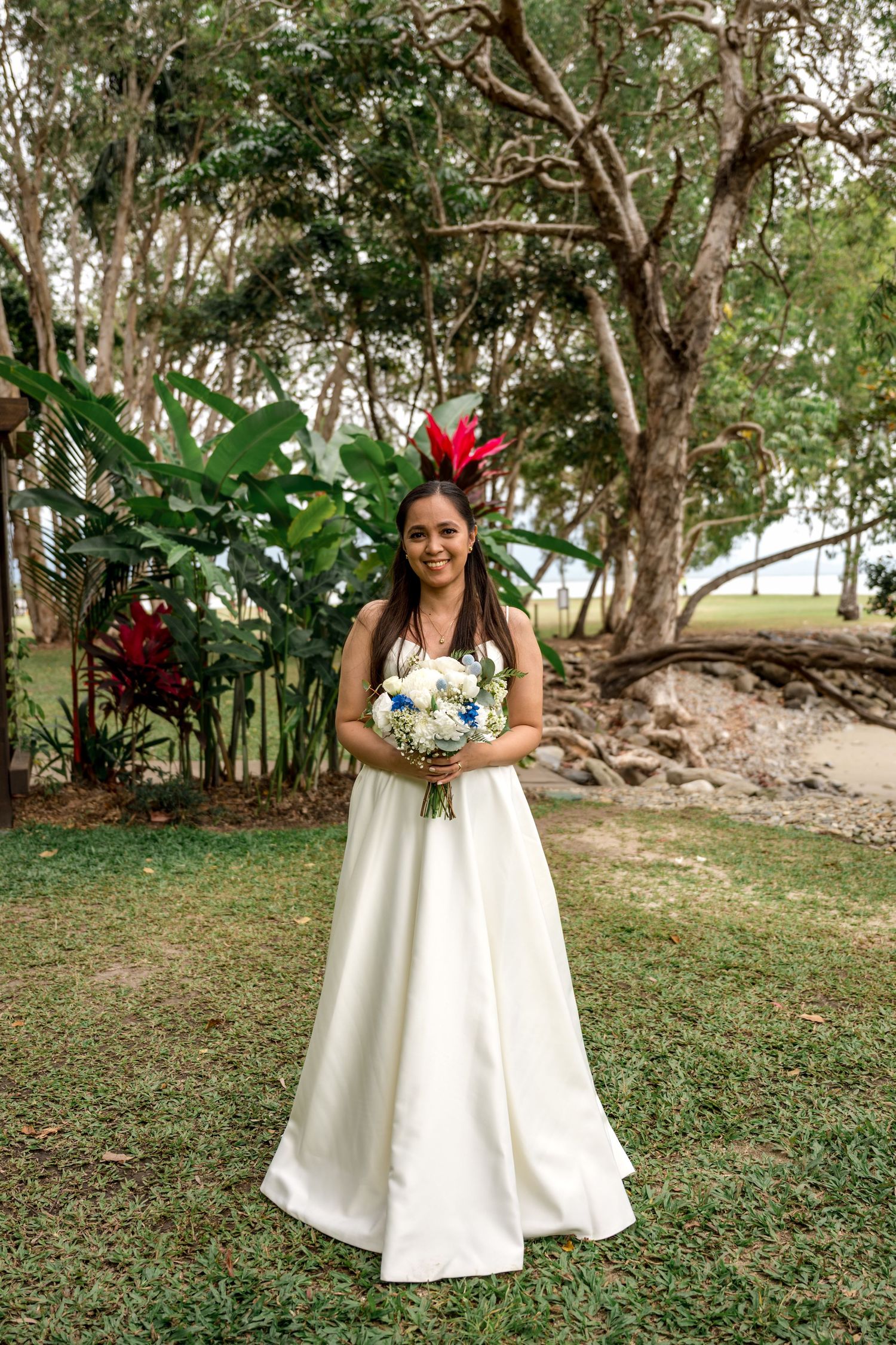 Smiling bride in white gown holds bouquet amid lush tropical garden greenery at Little Cove, Port Douglas.