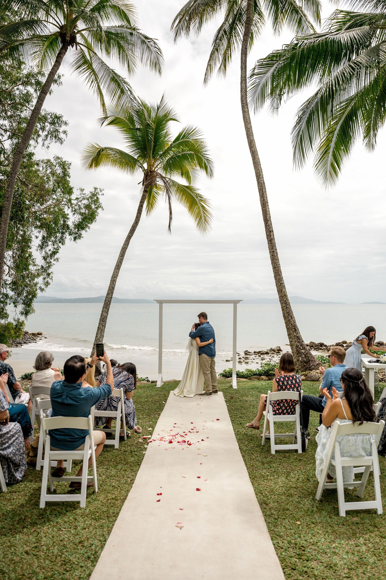 Beachside wedding ceremony under white arch flanked by tall palm trees with seated guests at Little Cove, Port Douglas.