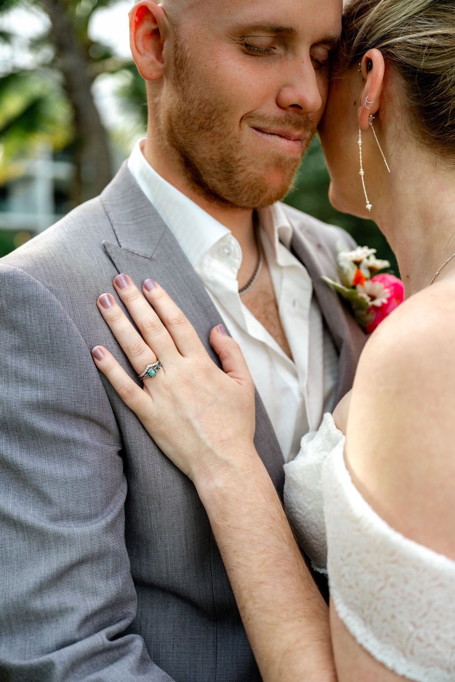 Close-up of bride resting hand on grooms grey suit jacket revealing emerald engagement ring with floral boutonniere.