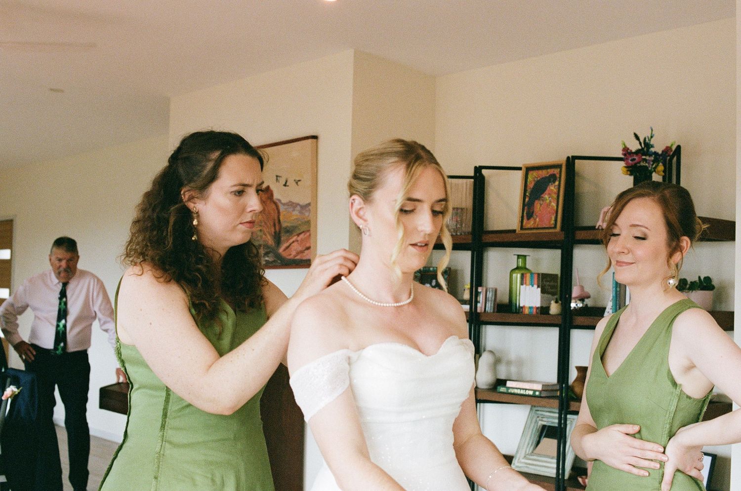 Bridesmaids in green dresses helping bride with pearl necklace during wedding preparations indoors.