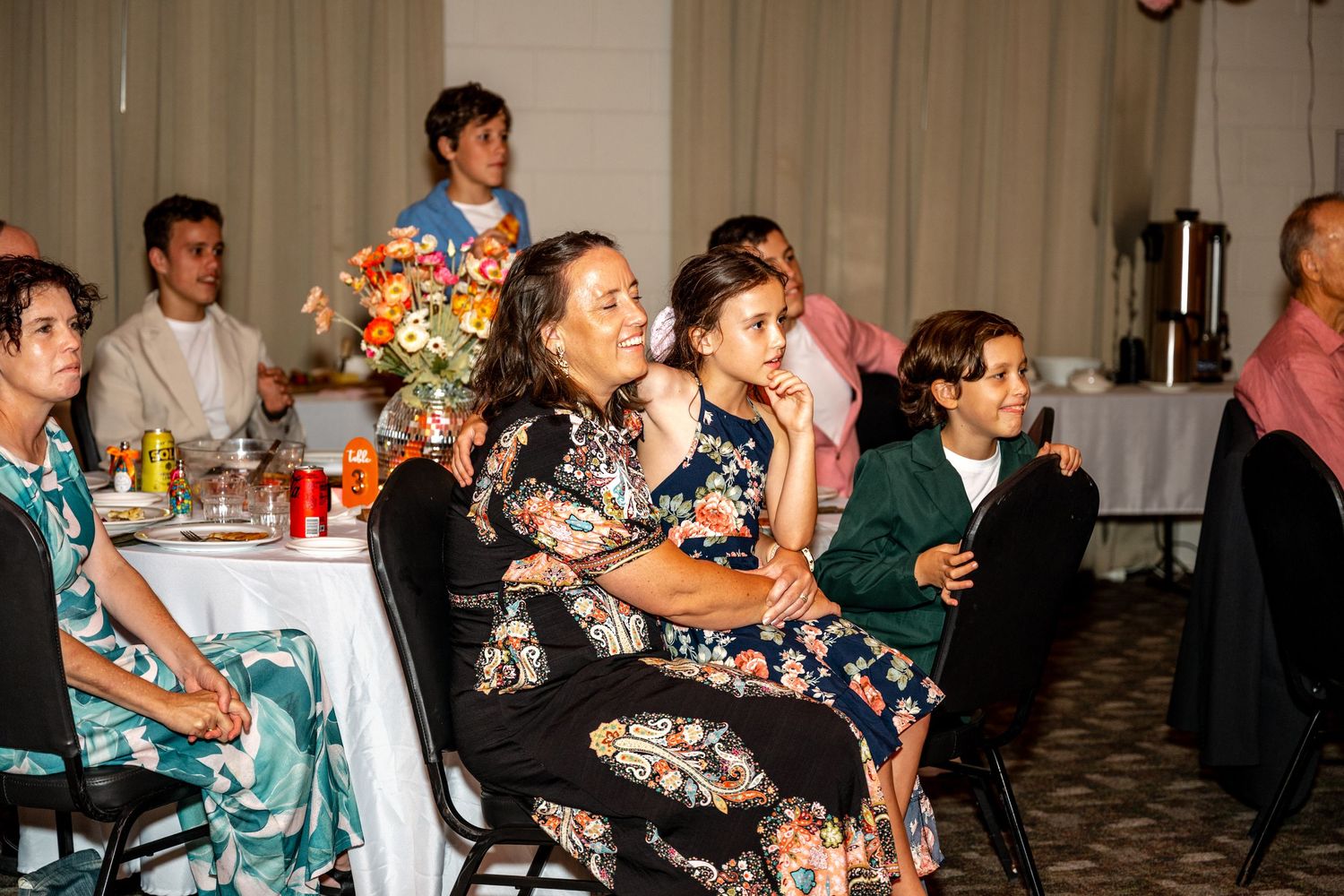 Wedding guests seated at reception tables watch attentively during indoor wedding reception celebration with floral centerpieces.