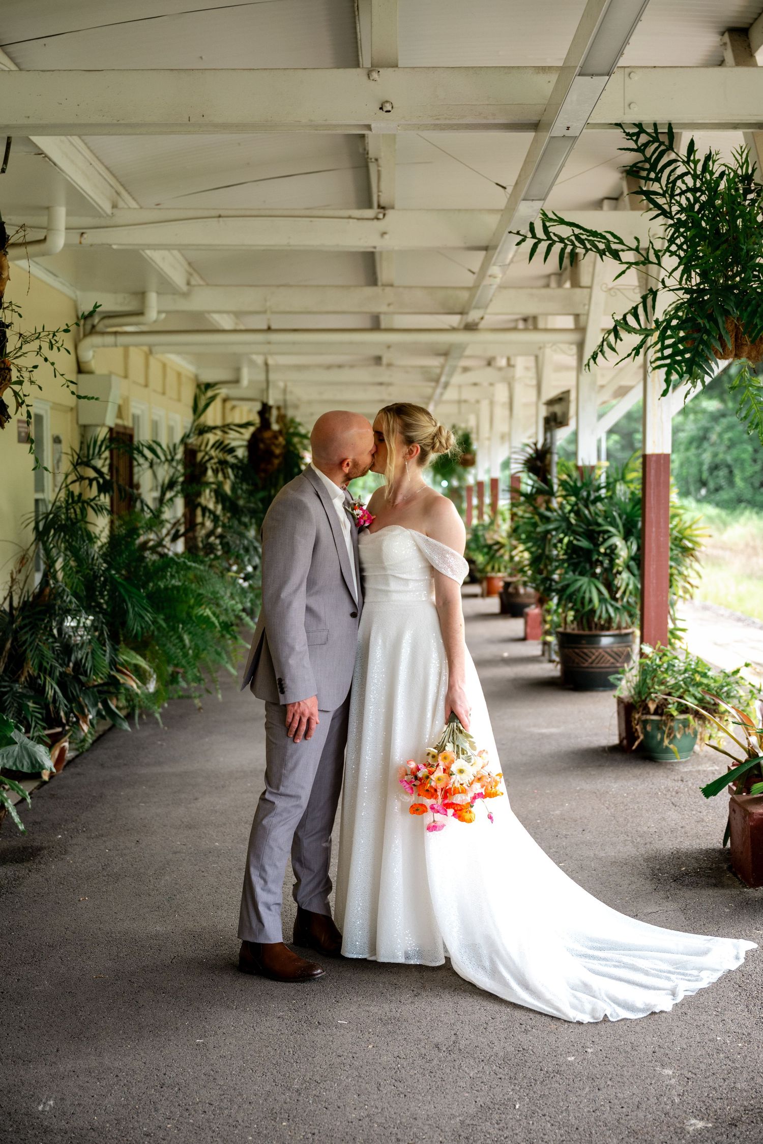 Bride and groom share a romantic kiss in a covered tropical venue corridor lined with lush plants.