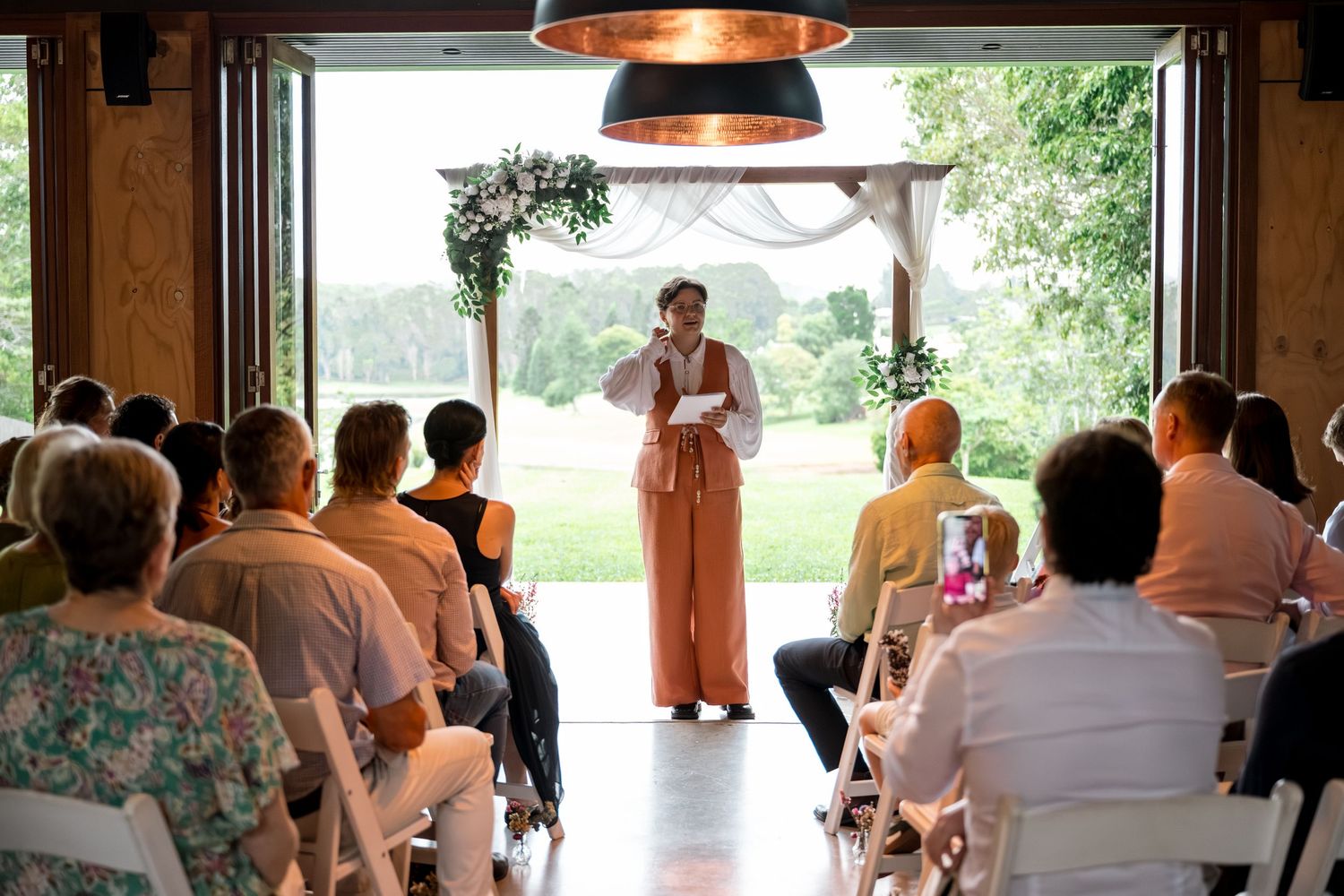 An officiant in a peach dress addresses wedding guests seated in a bright open barn venue.